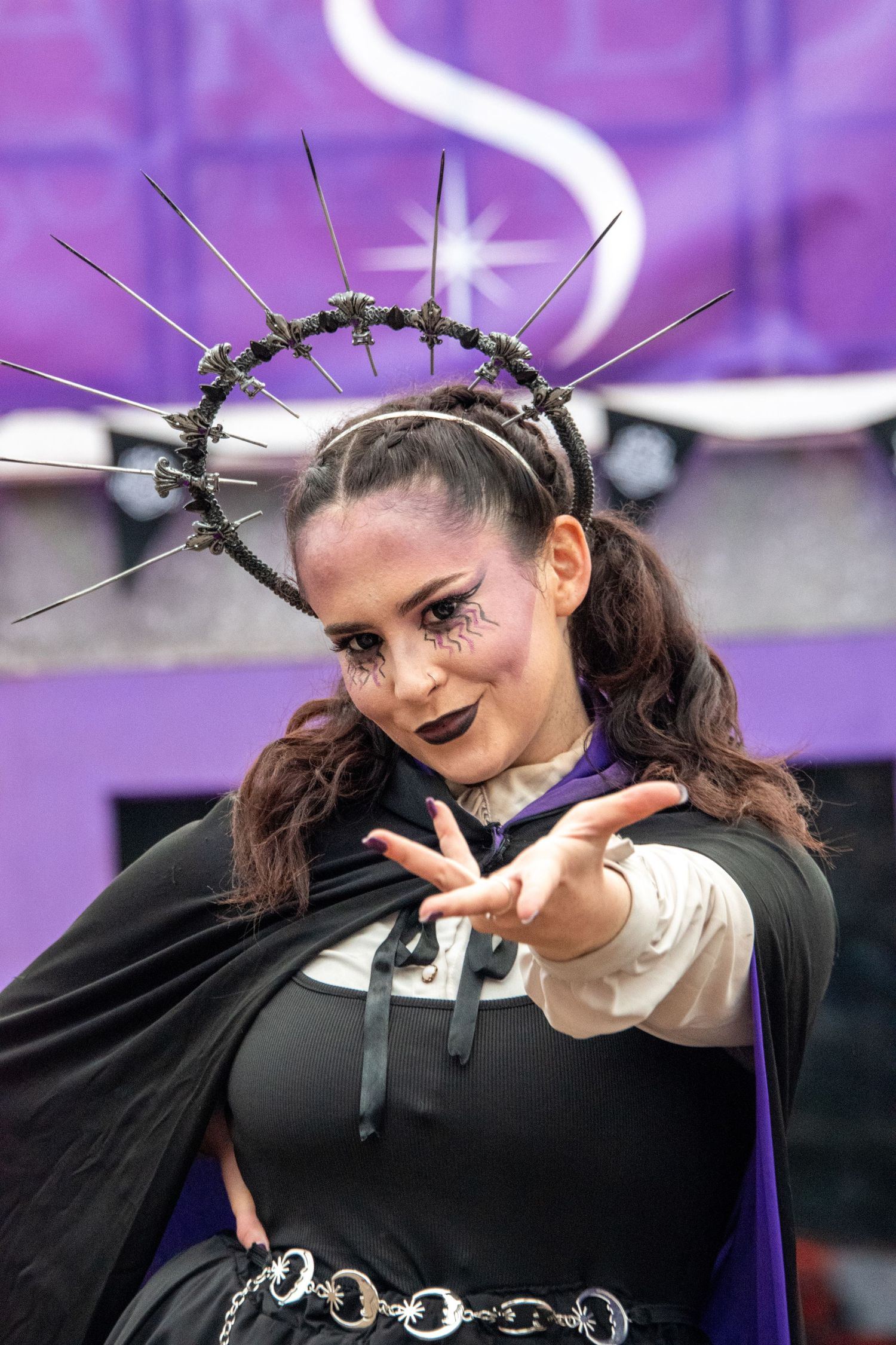 Woman in gothic black costume with spiked halo headpiece reaches toward camera dramatically.