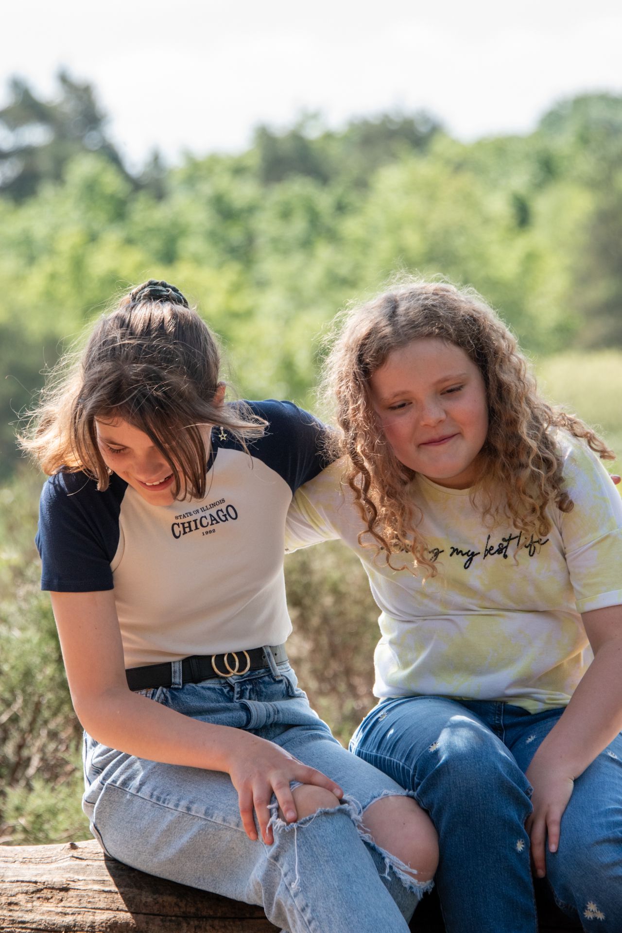 Two young girls sit together on a log outdoors, smiling in casual jeans and graphic tees.