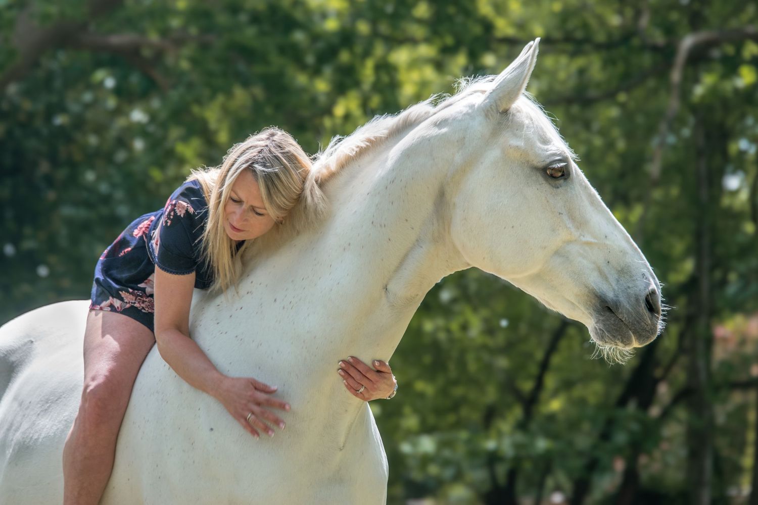 Blonde woman leaning forward embracing a white horse outdoors.