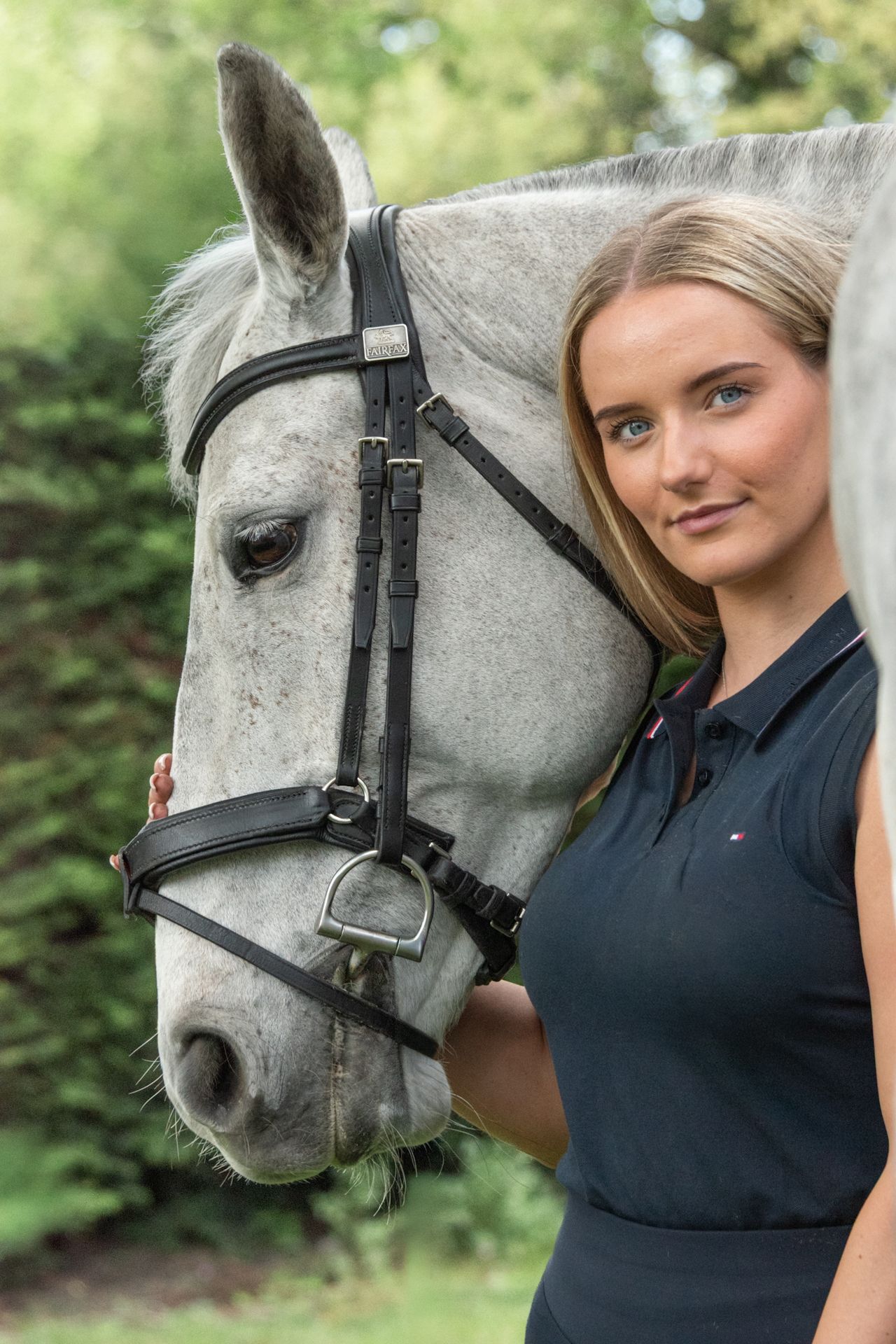 Young blonde woman in navy polo shirt posing with a grey horse wearing a black bridle outdoors.