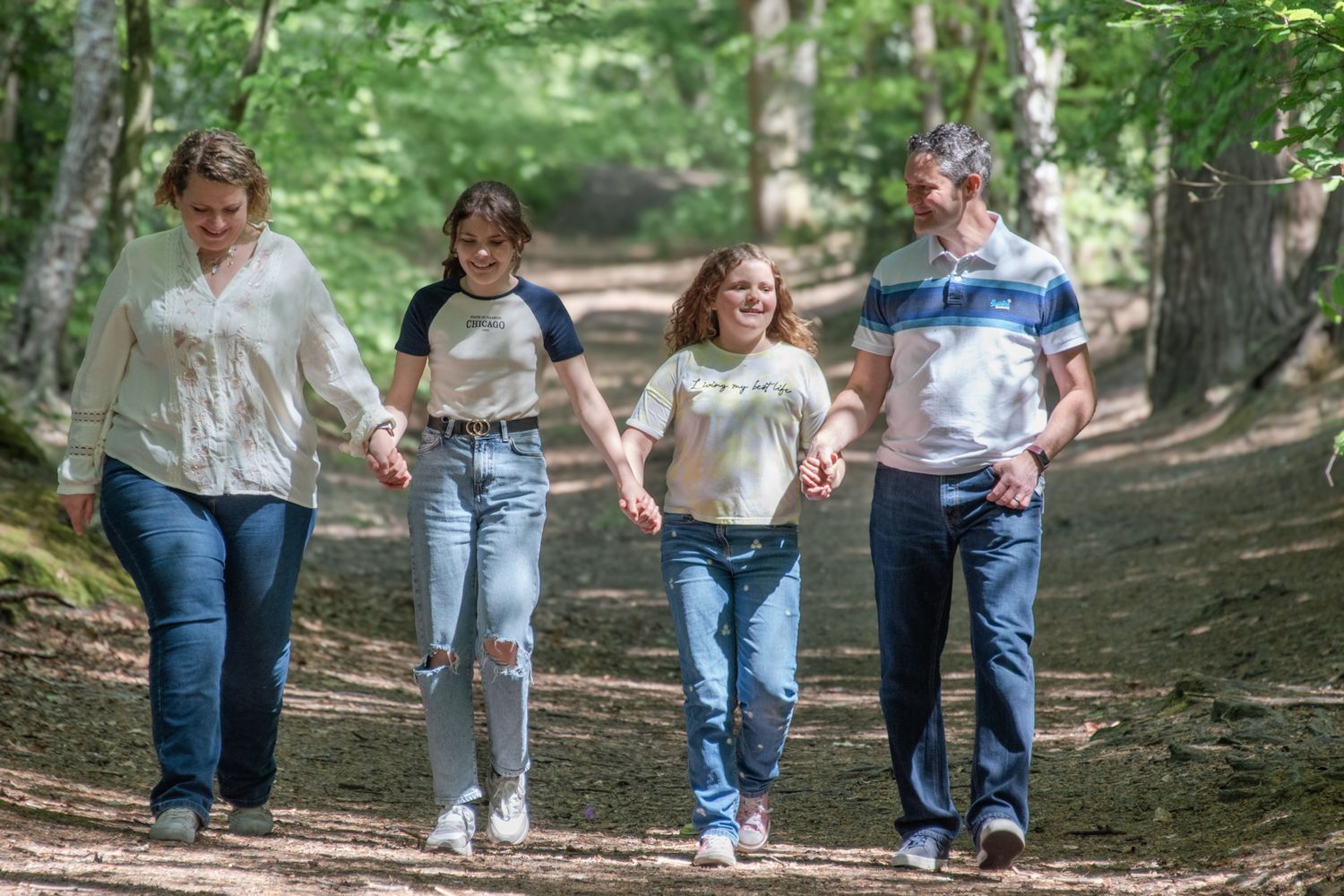 Family of four holding hands walking along a sunlit woodland path surrounded by trees.