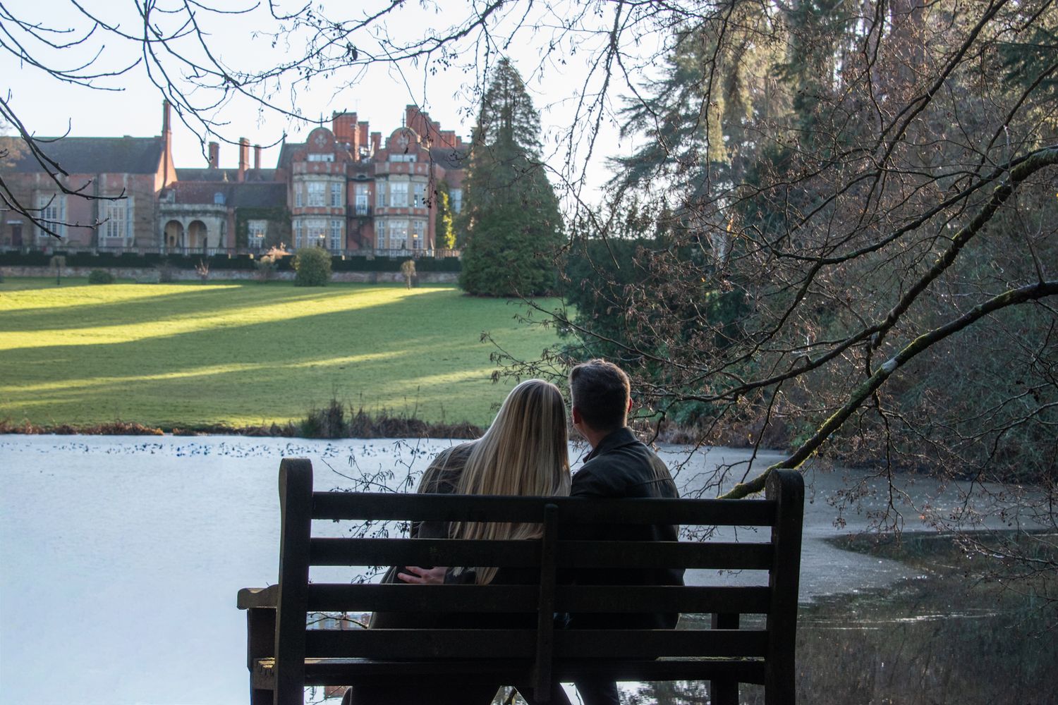 Engaged couple sitting on a bench overlooking a lake with a grand red brick manor house behind them.