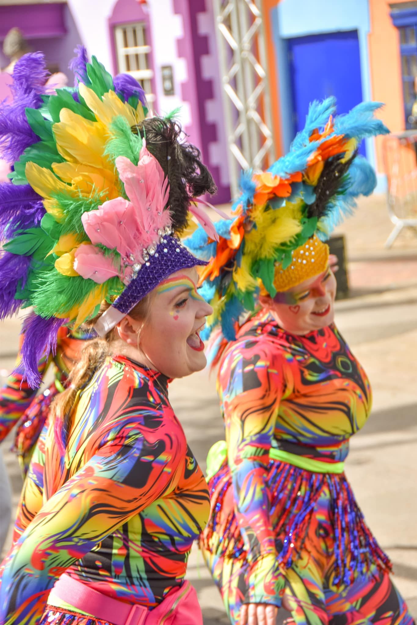 Two women in bright multicolored costumes with feathered headdresses perform at a lively carnival parade.