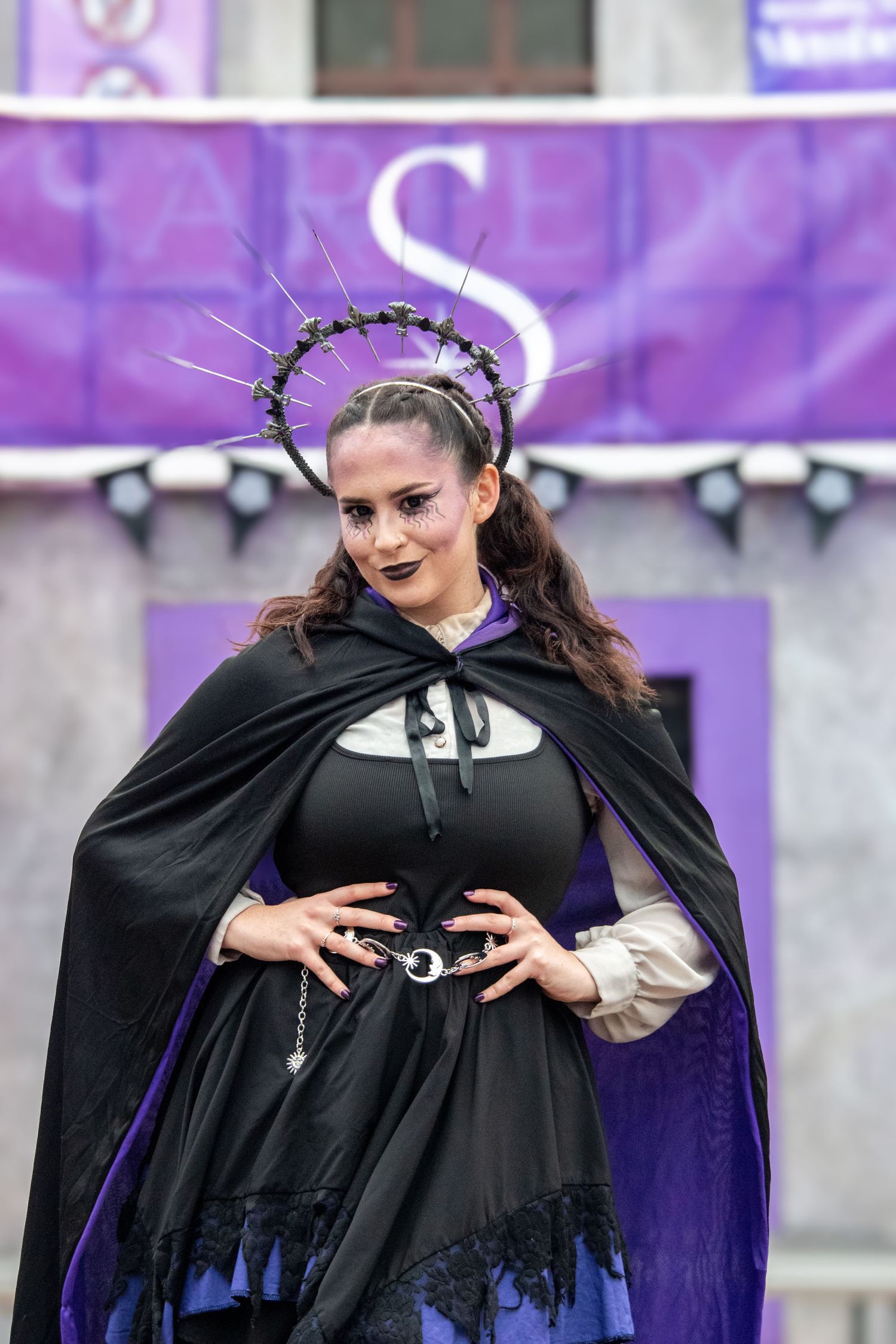 Woman in gothic black cape and spiked crown headpiece poses in dramatic Halloween costume.