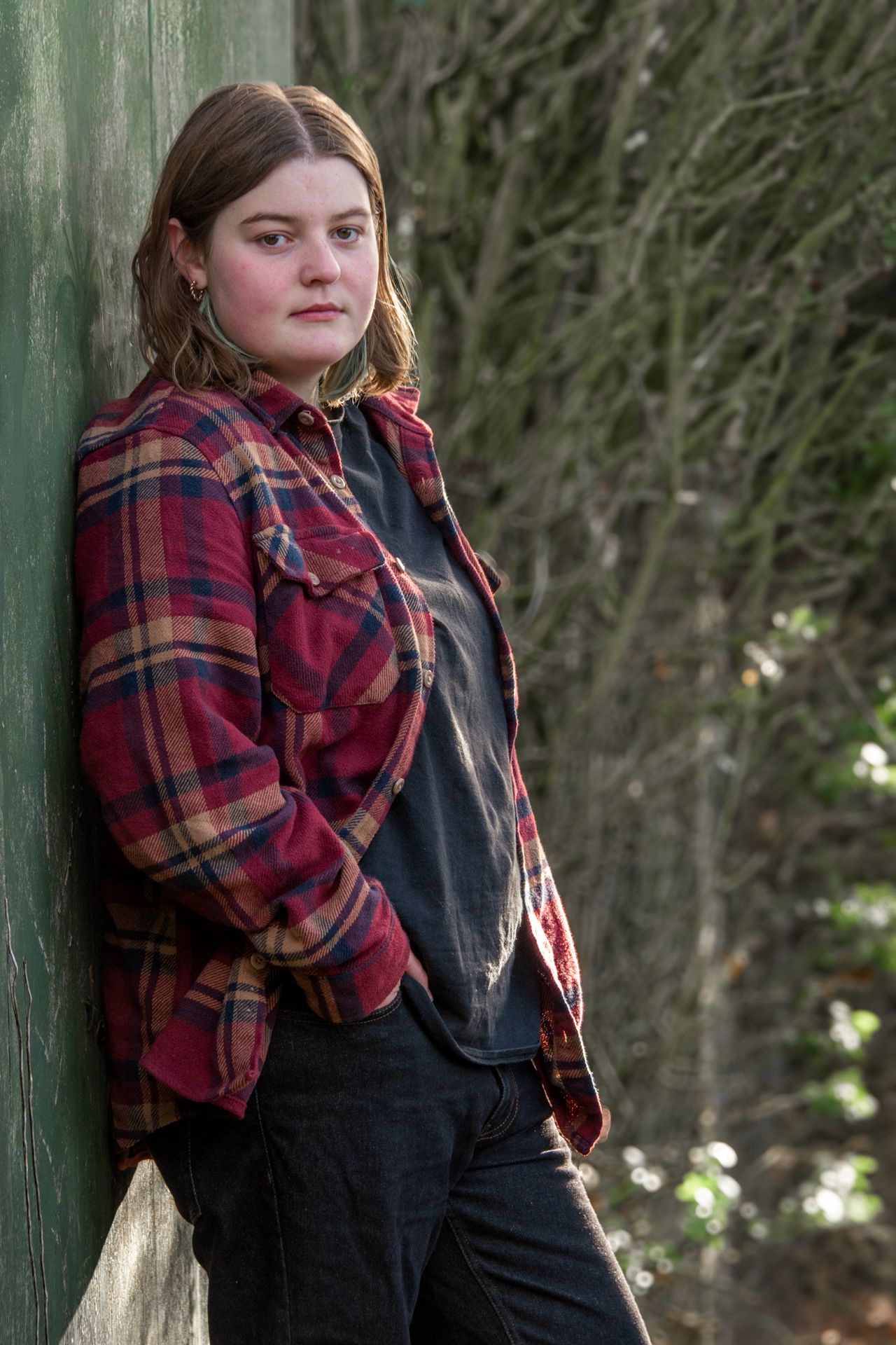 Teen girl in red plaid shirt leaning against green post with serious expression in woodland setting.
