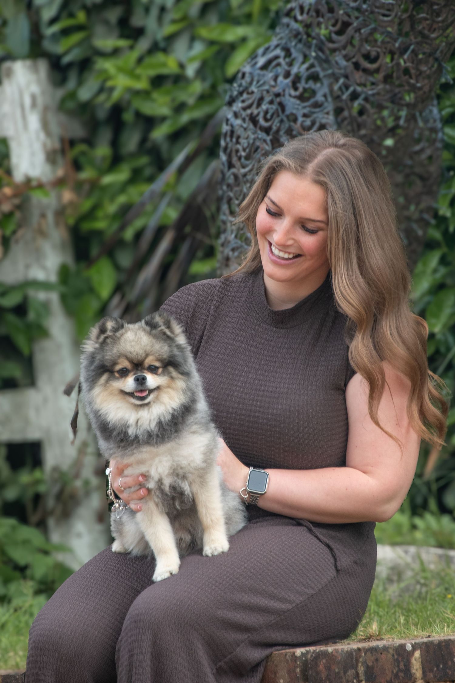 Smiling young woman in a brown outfit sitting outdoors holding a small Pomeranian dog.