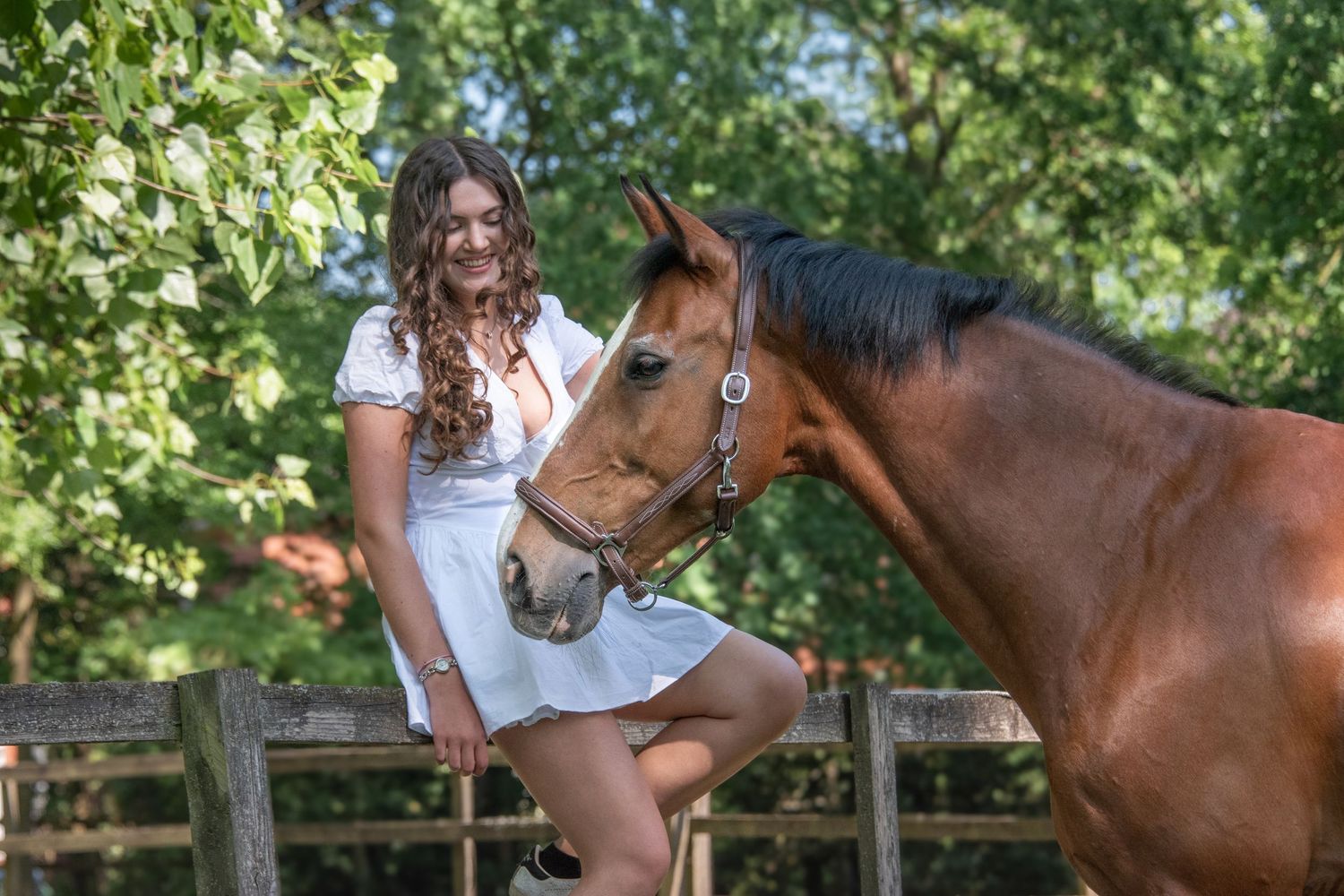 A smiling young woman in a white dress sits on a fence beside a bay horse in a sunny paddock.