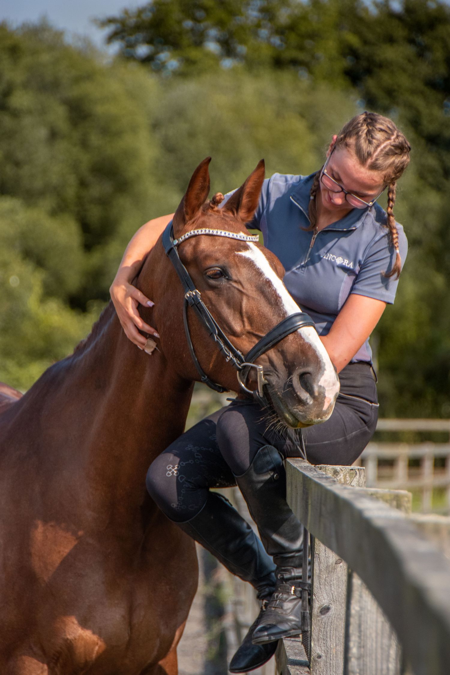 A woman with braids leans over a fence to affectionately pet a chestnut horse outdoors.