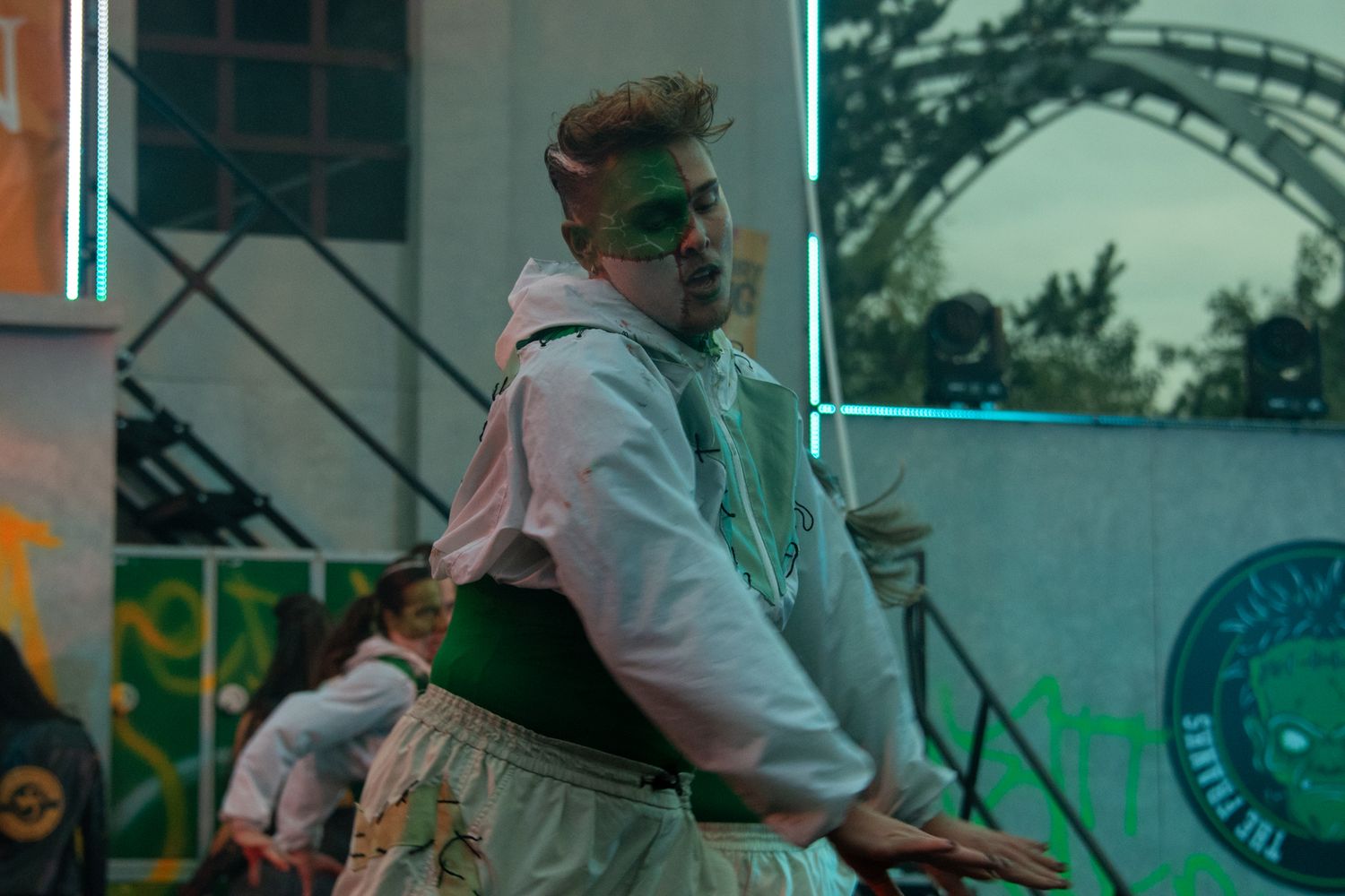 Male performer with green face paint dancing in a white costume under colorful stage lights at an outdoor event.