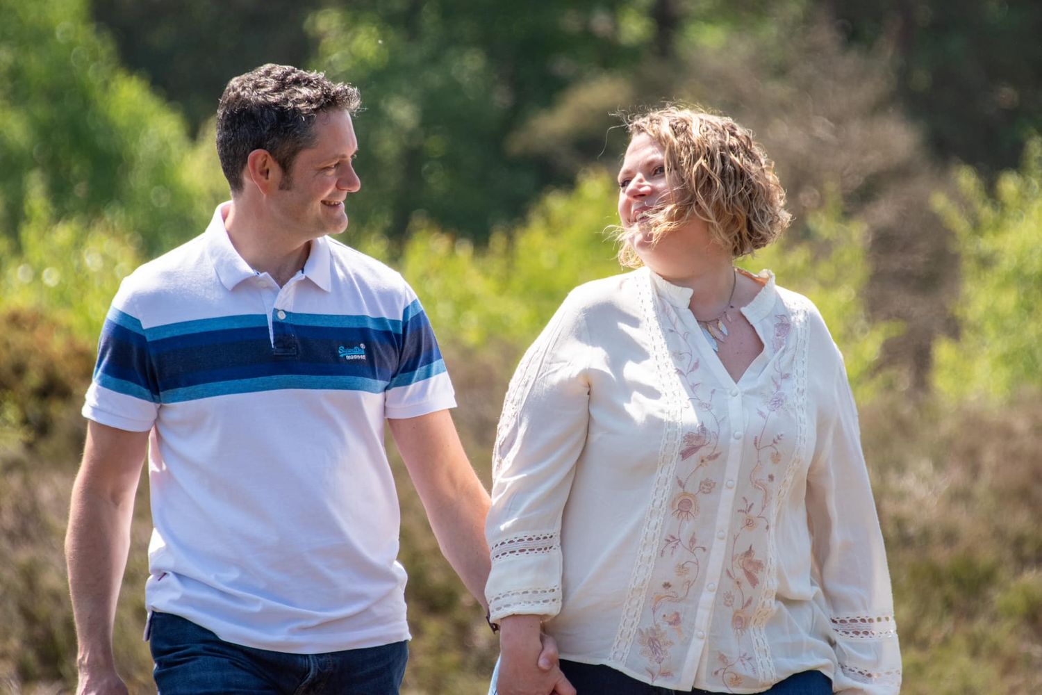 Happy couple walking hand in hand outdoors, smiling at each other in a bright sunlit natural setting.