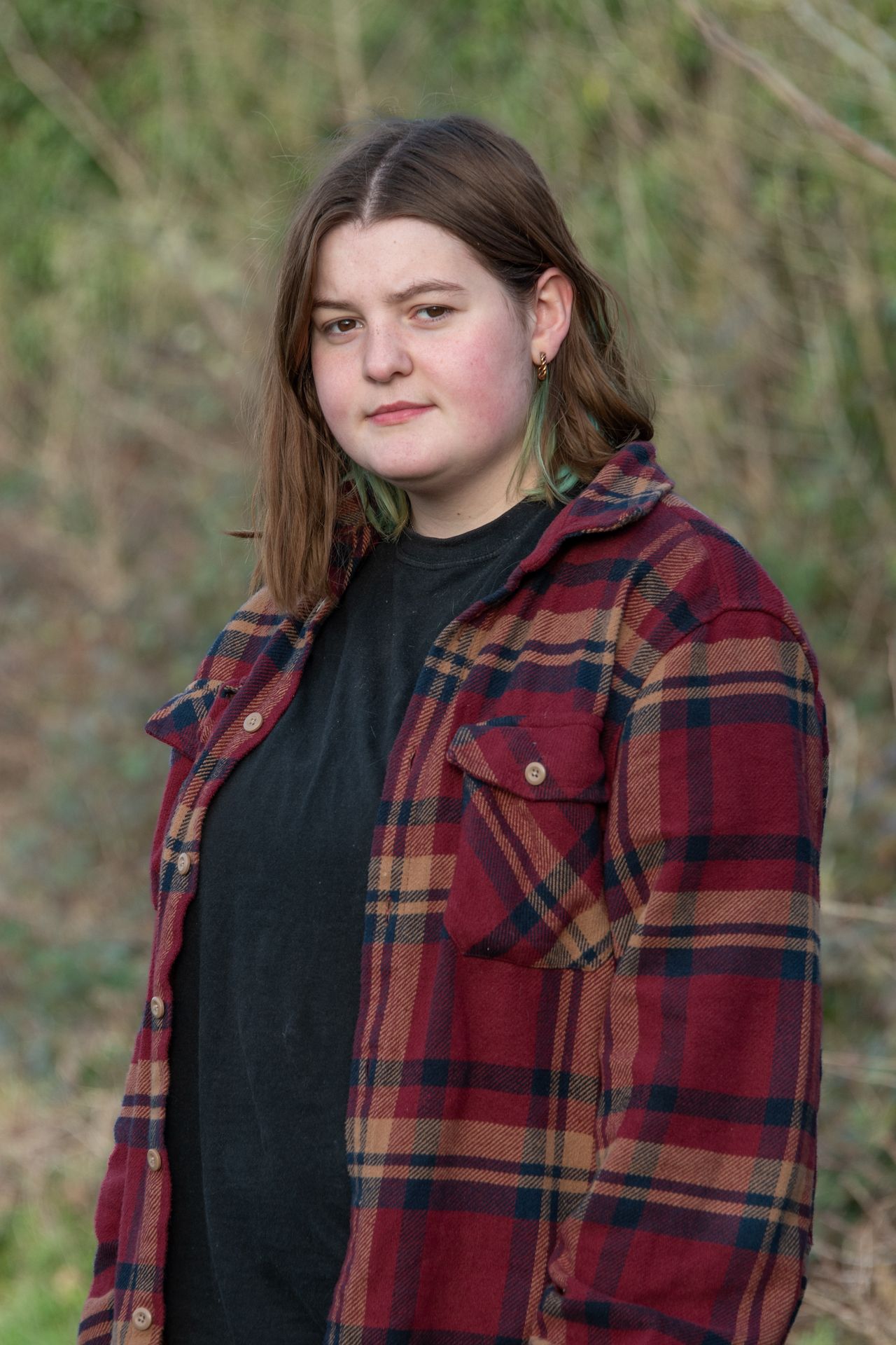 Young woman wearing red plaid flannel shirt standing outdoors with natural greenery background.