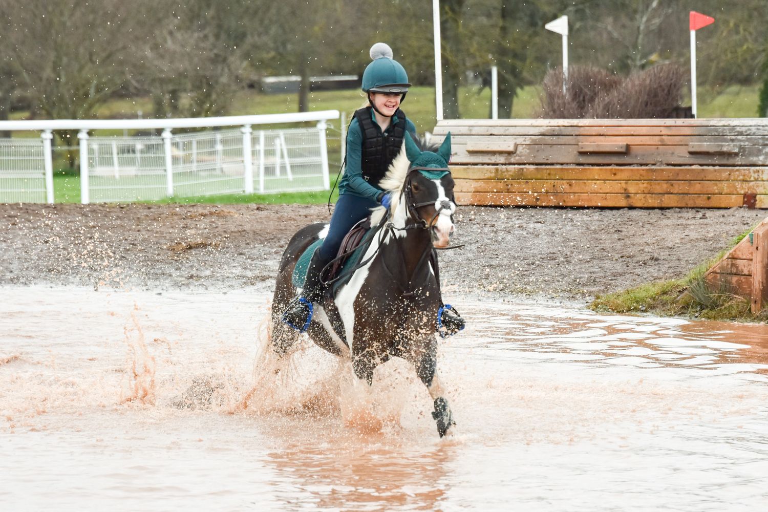 Rider on a black and white horse splashing through water during an equestrian cross-country event.