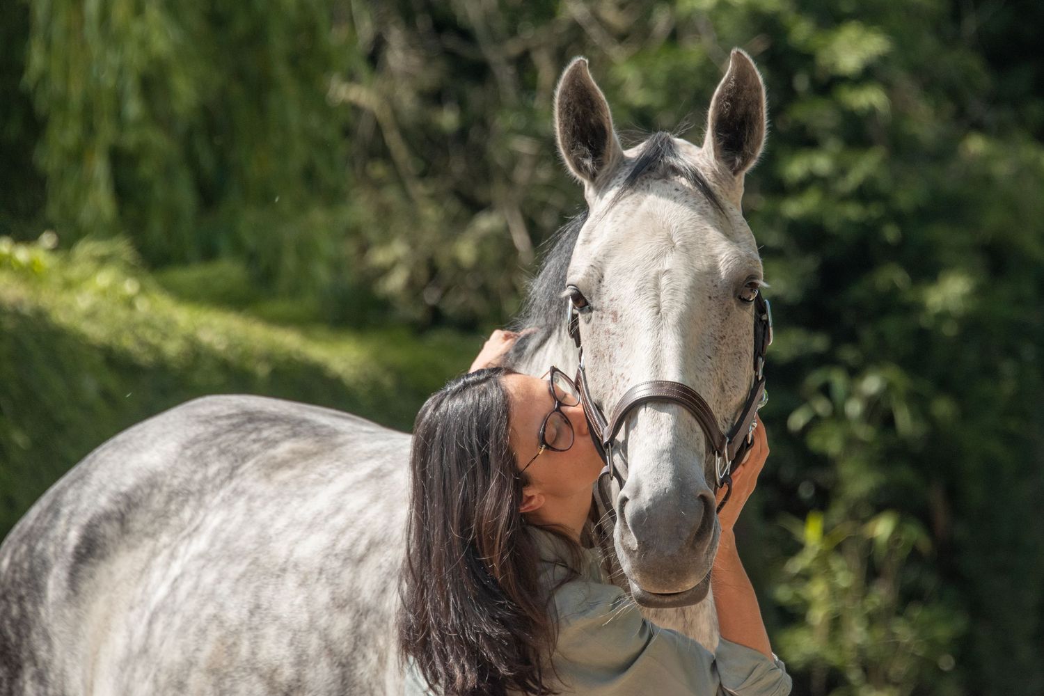 Woman kissing a dapple gray horse wearing a bridle outdoors with lush green trees in the background.