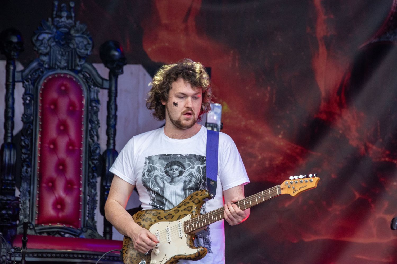 Male guitarist in white t-shirt playing a gold electric guitar on an outdoor festival stage.