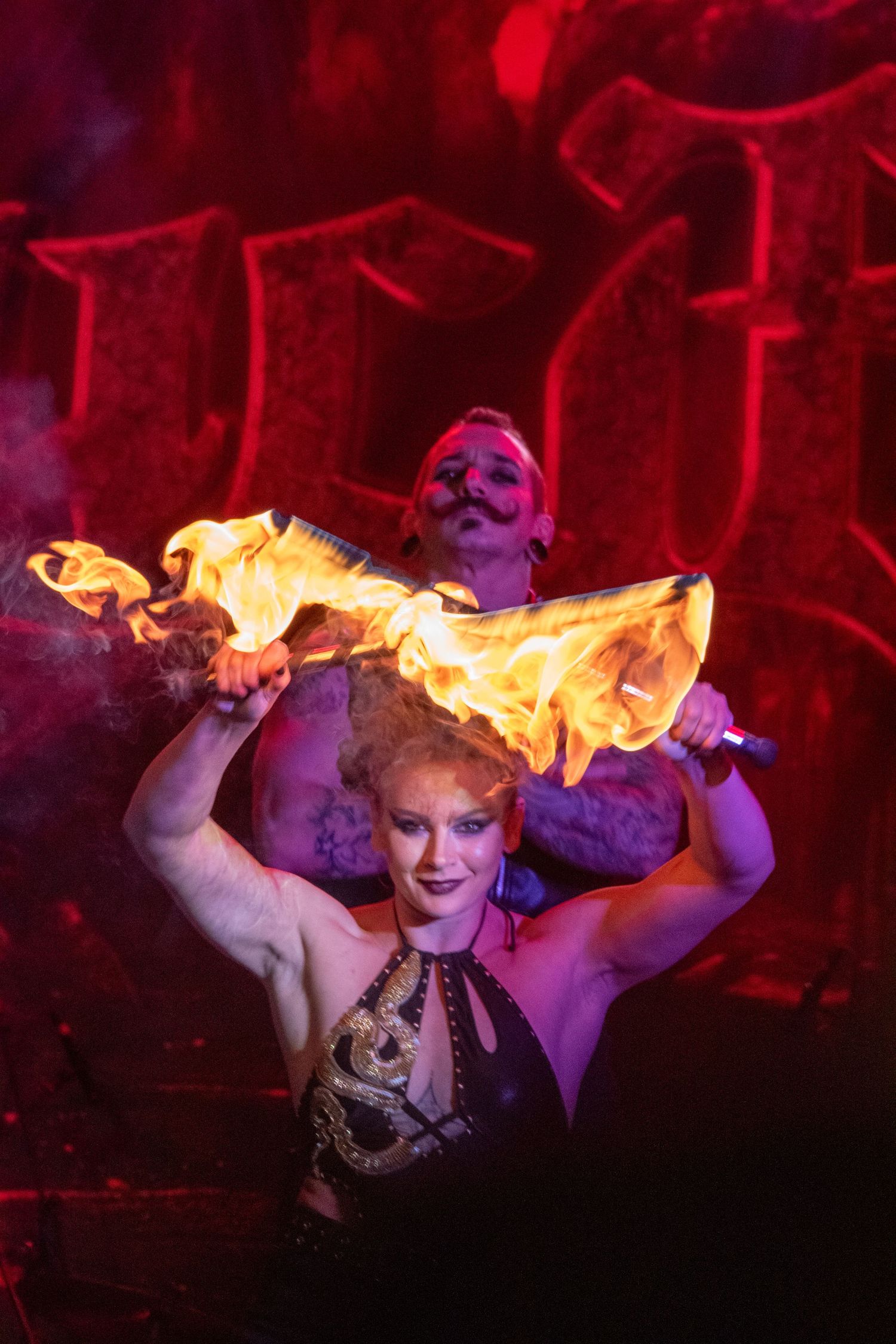 Fire performer holding flaming torches above her head on a red-lit stage with male performer.
