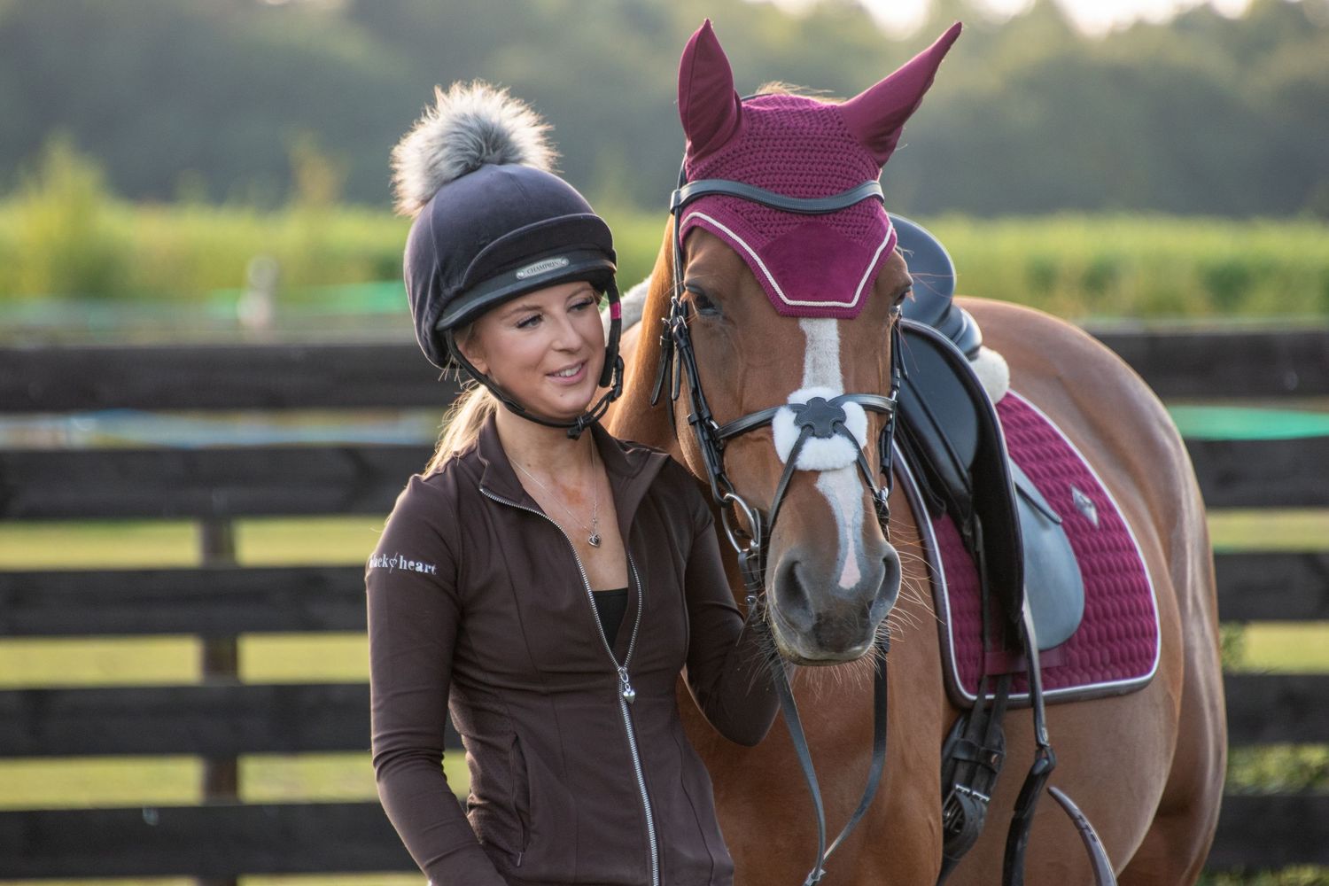 Smiling woman in riding hat standing beside a chestnut horse outdoors.