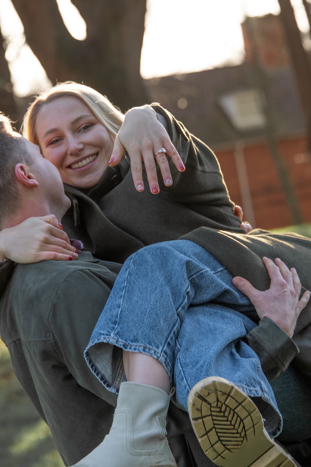 Smiling blonde woman being carried showing engagement ring while laughing outdoors.