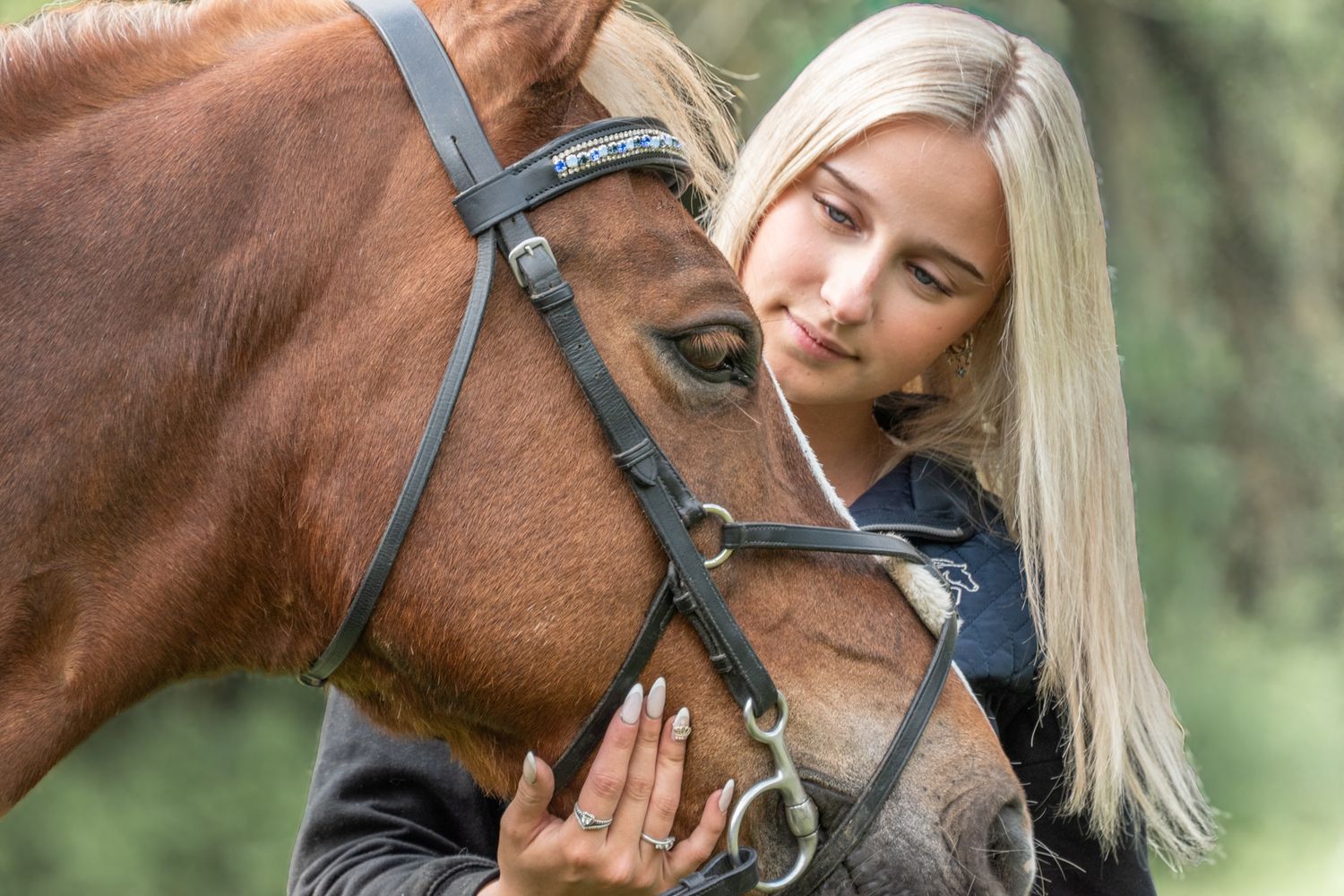 Blonde woman gently pressing her face against a brown horse wearing a black bridle outdoors.