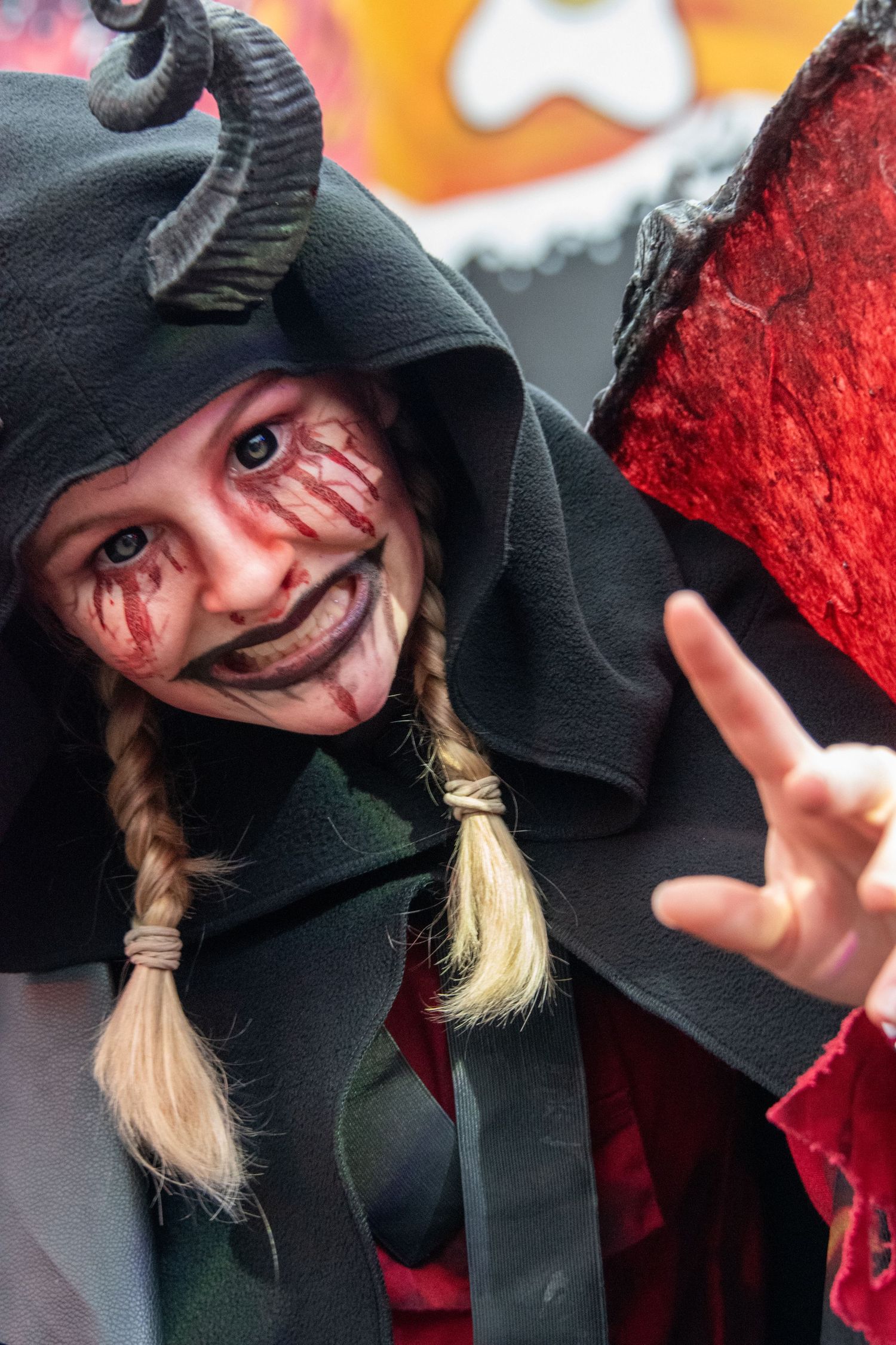 Child in dark devil Halloween costume with horns, bloody face makeup, braids, making a hand gesture.