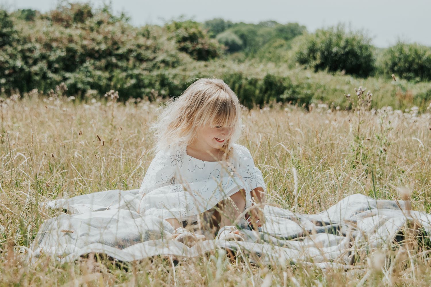 Young blonde child in white dress playing on a blanket in a tall grass meadow.