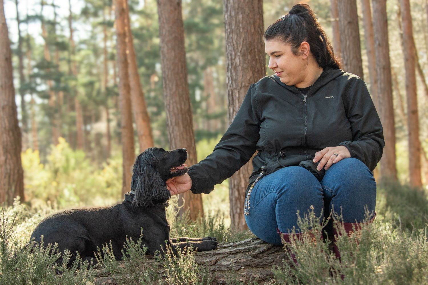Woman in black jacket trains black Cocker Spaniel dog in a pine forest setting.