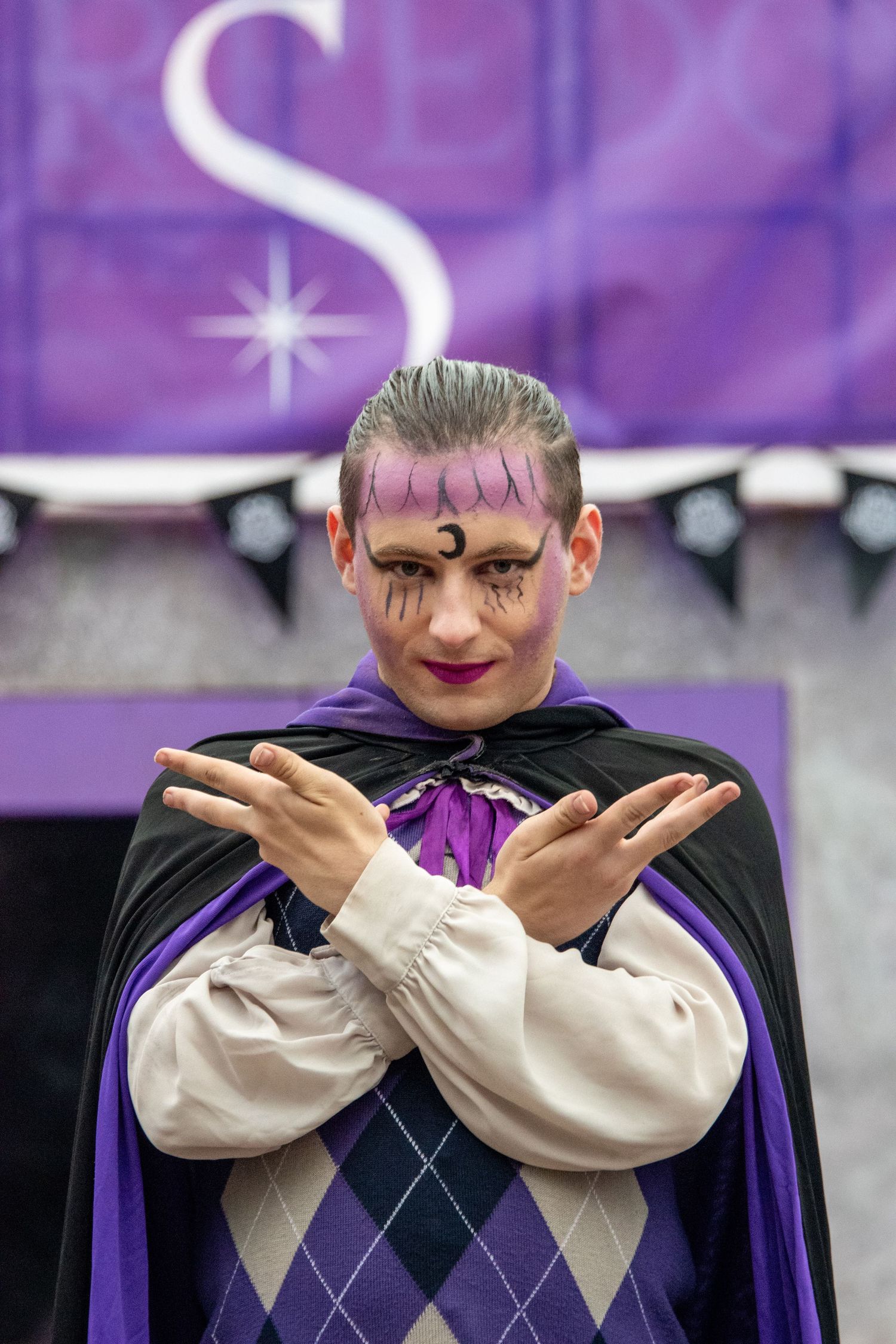 Performer in purple argyle vest and black cape with dramatic face paint crosses arms on stage.