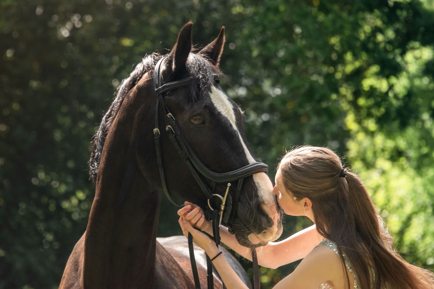 Young girl kissing a dark brown horse wearing a bridle outdoors.