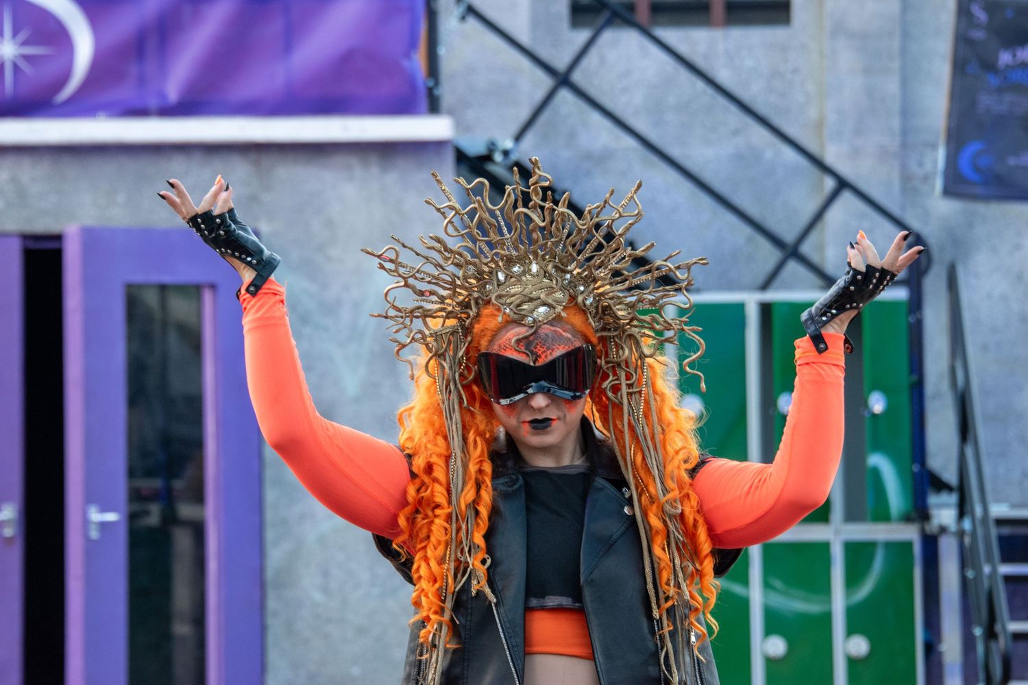 Performer in orange outfit with elaborate golden headdress raises arms on outdoor stage.