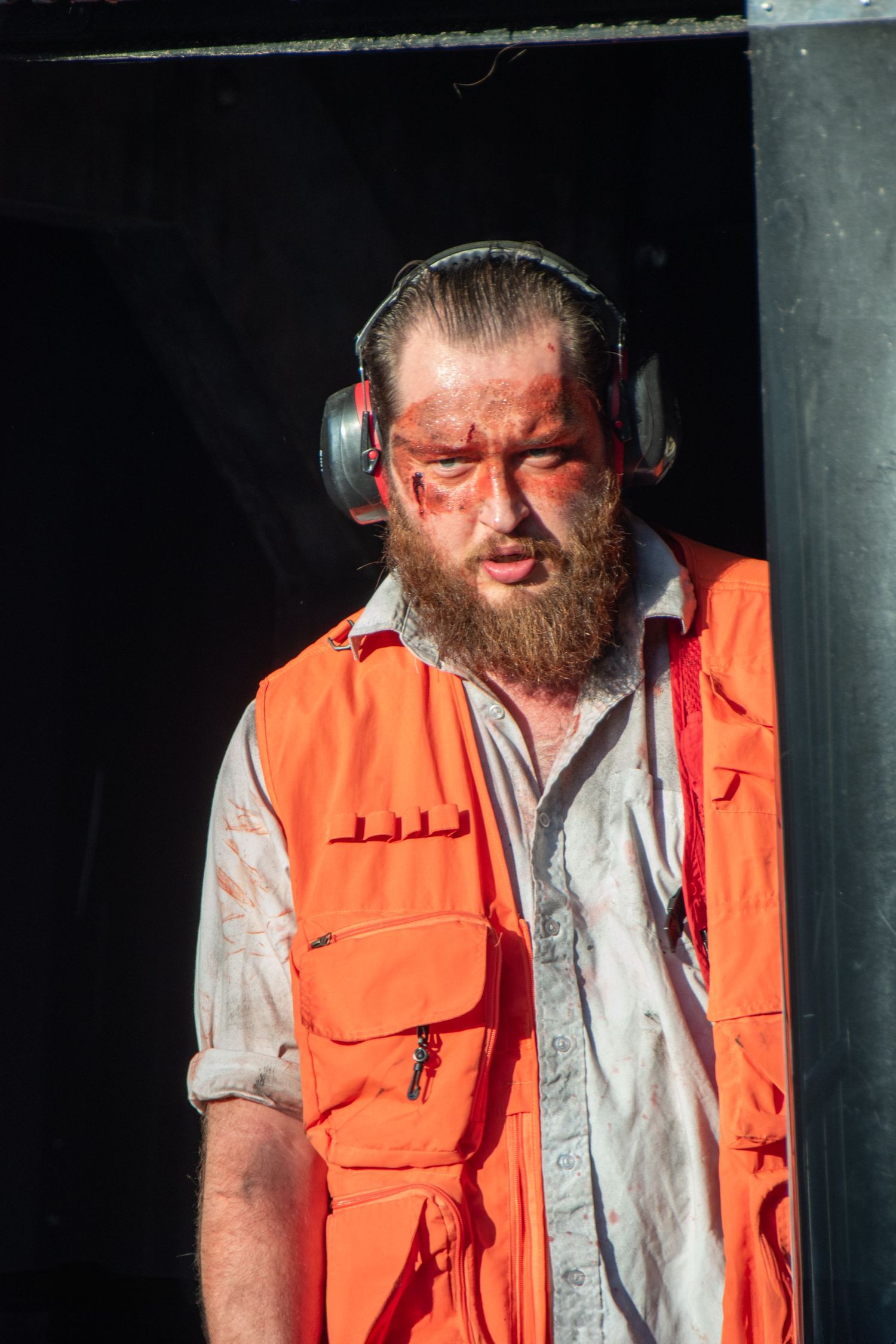 Bearded man in horror costume with bloody face makeup, orange vest, headphones, standing in dark doorway.