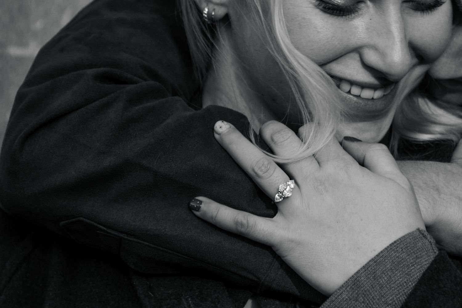 Close-up of smiling woman showing diamond engagement ring while embracing partner in black and white.