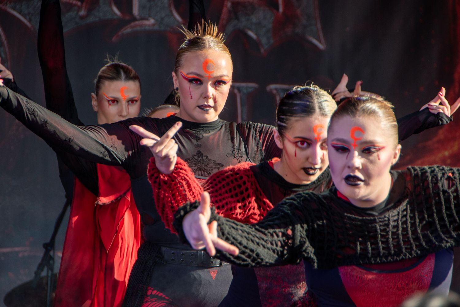 Group of female dancers in dark costumes with red and black face paint performing on stage.