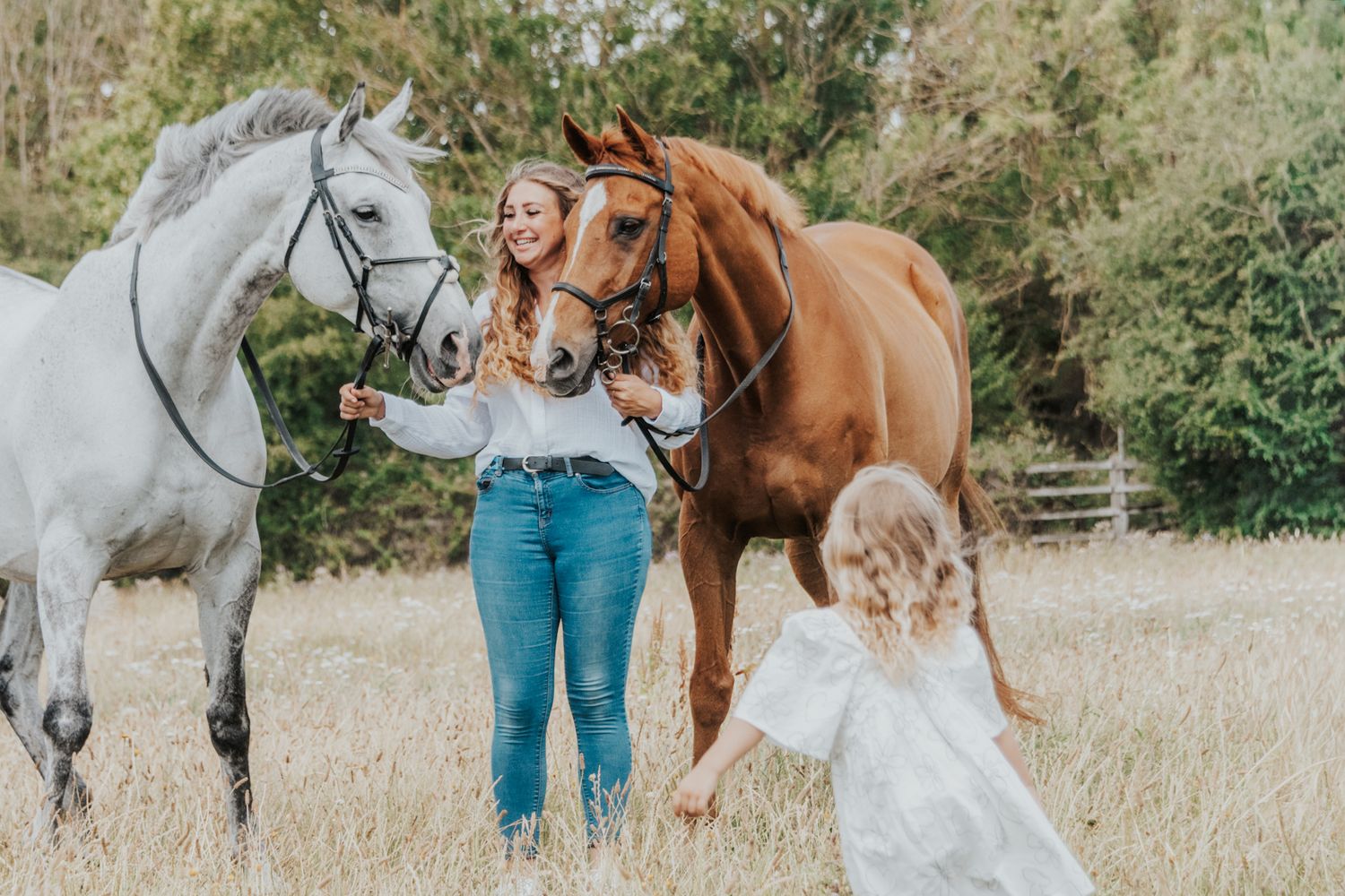 Woman holding two horses, a white and chestnut, in a field while a young girl in a white dress approaches.