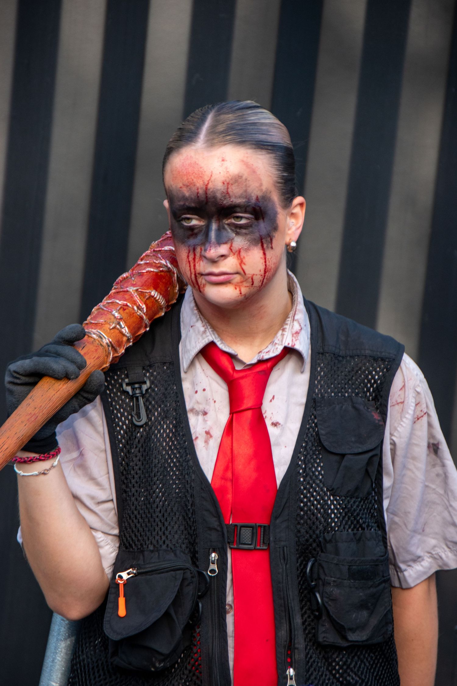 Person in zombie costume with dark face paint, blood effects, red tie, and barbed wire bat prop.
