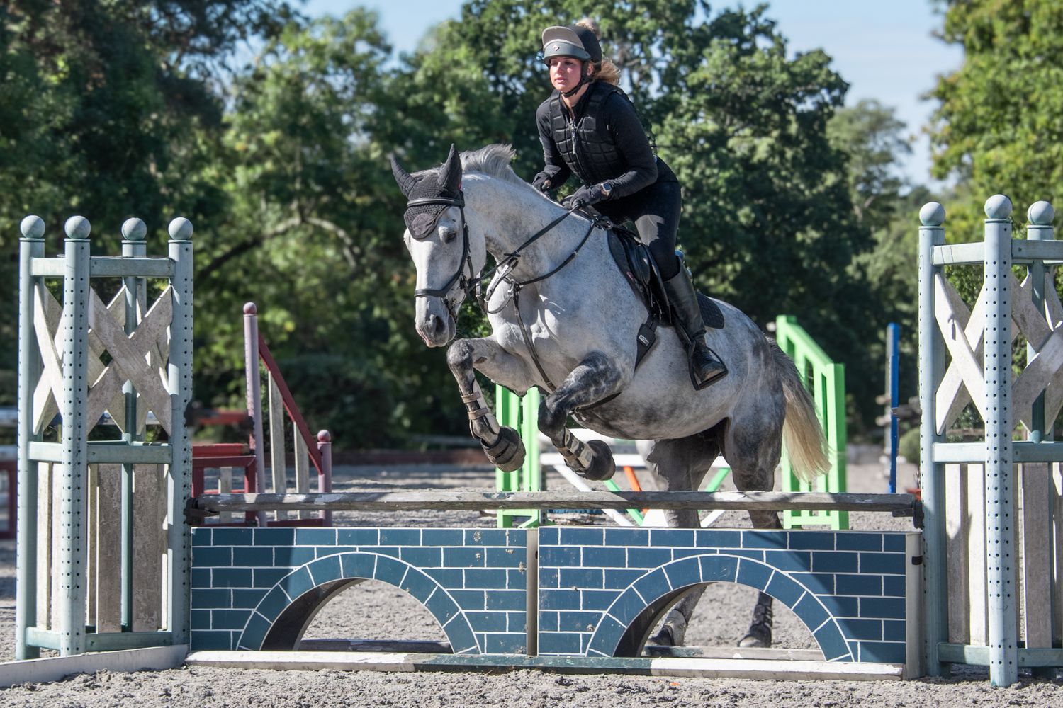 Equestrian rider on a grey horse jumping over a decorative brick fence obstacle in a competition arena.
