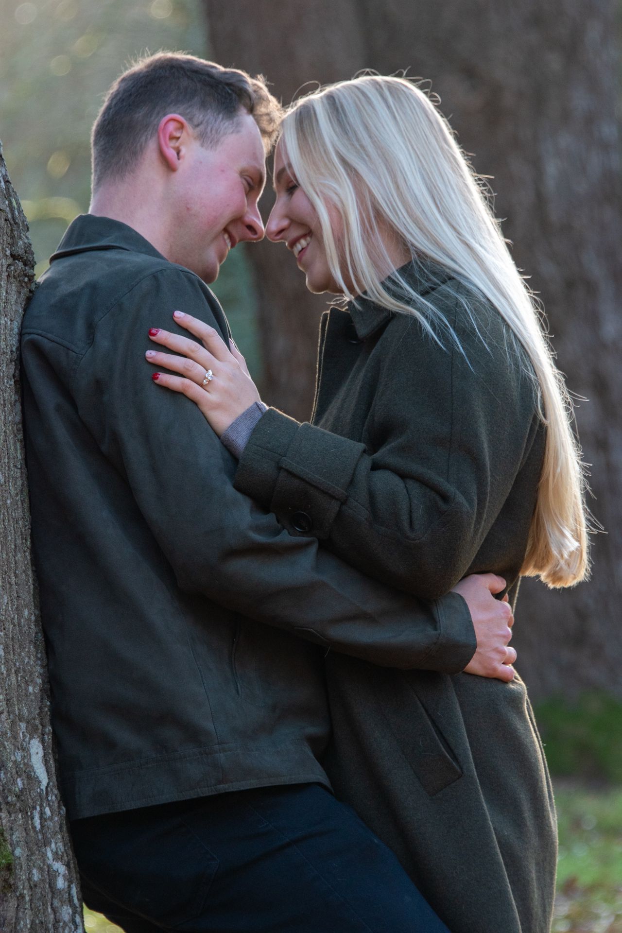 Smiling engaged couple leaning against a tree, touching foreheads in a tender romantic moment outdoors.
