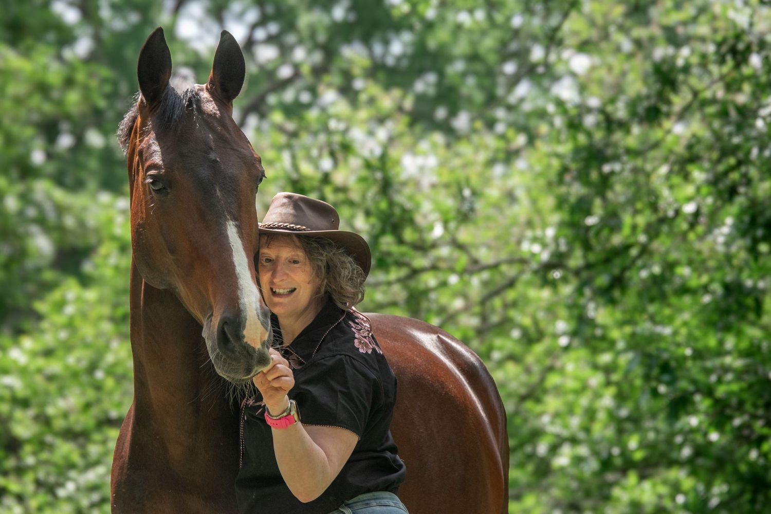 Smiling woman in a cowboy hat affectionately embracing a chestnut horse outdoors among green trees.