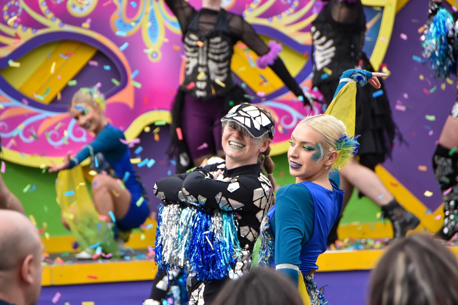 Performers in colorful skeleton and carnival costumes dance energetically during a festive parade celebration.
