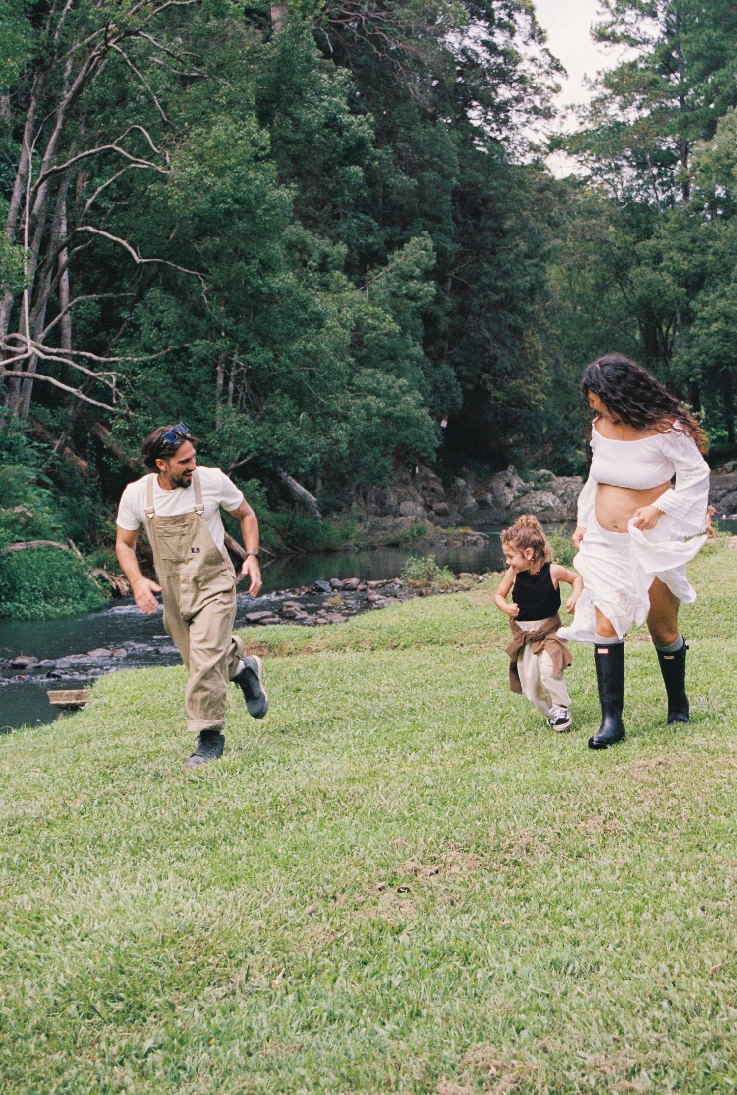 Family playing outdoors on green grass near a forest stream, with parents chasing a young child.