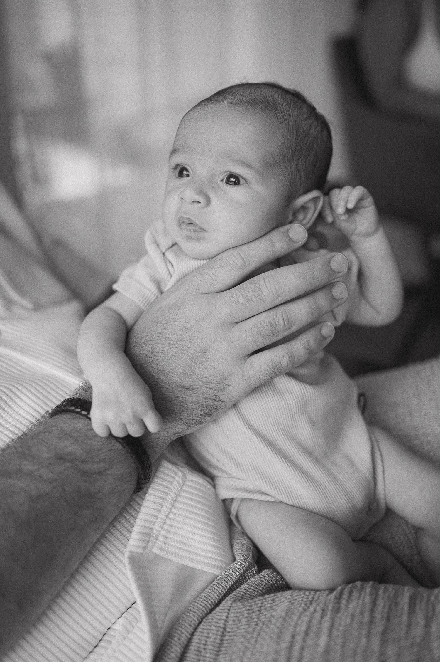 Black and white photo of a newborn baby being cradled in an adult's hands, gazing upward with curious eyes.
