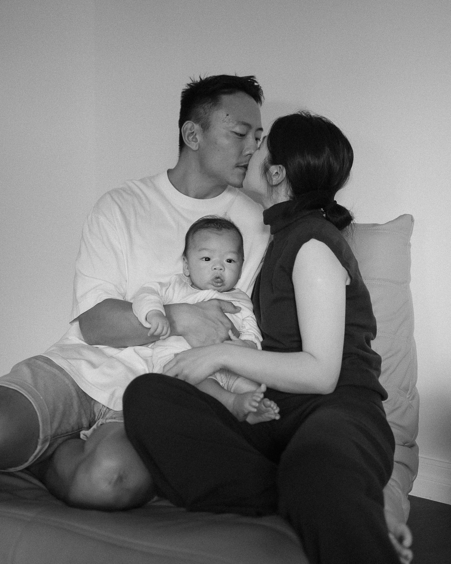 Black and white photo of a couple sharing a kiss while holding their newborn baby on a couch.