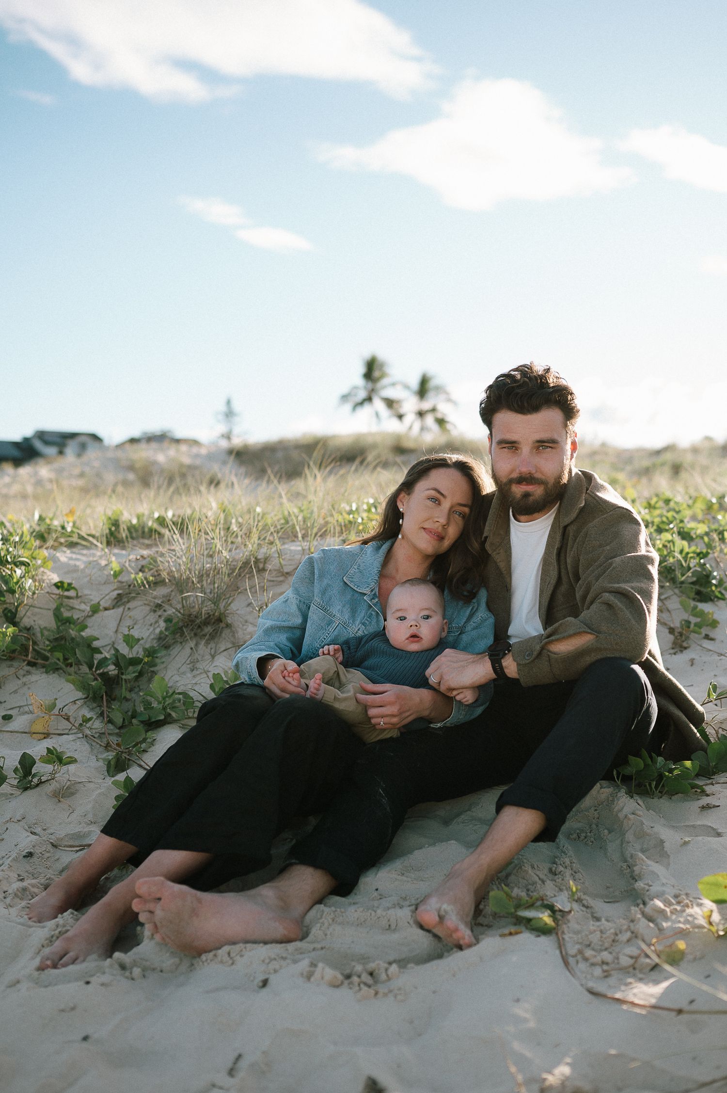 Family relaxing together on sandy dunes with palm trees and blue sky in the background.
