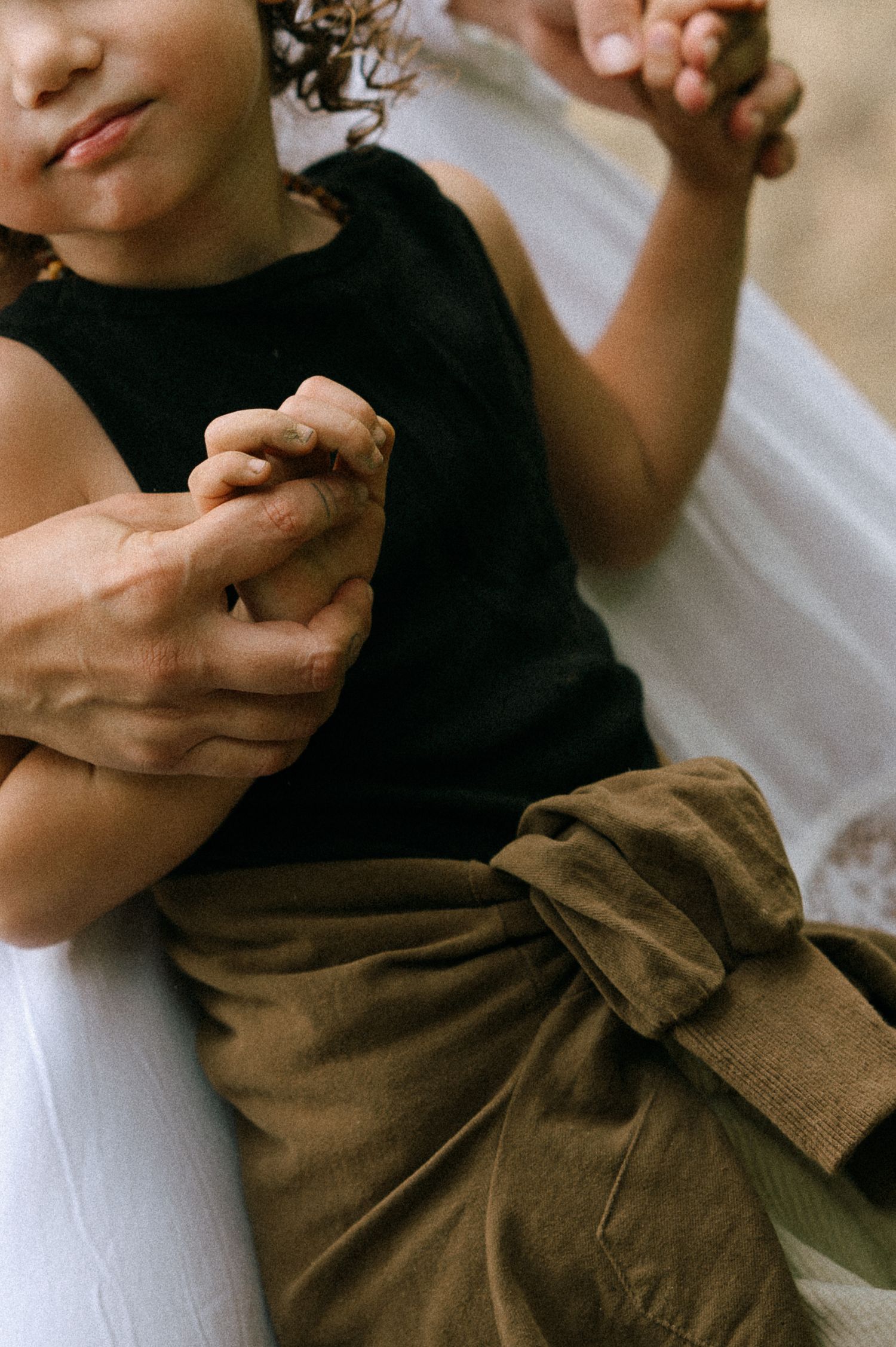 Adult holding a child's hands tenderly, child wearing black top and olive green tied pants outdoors.