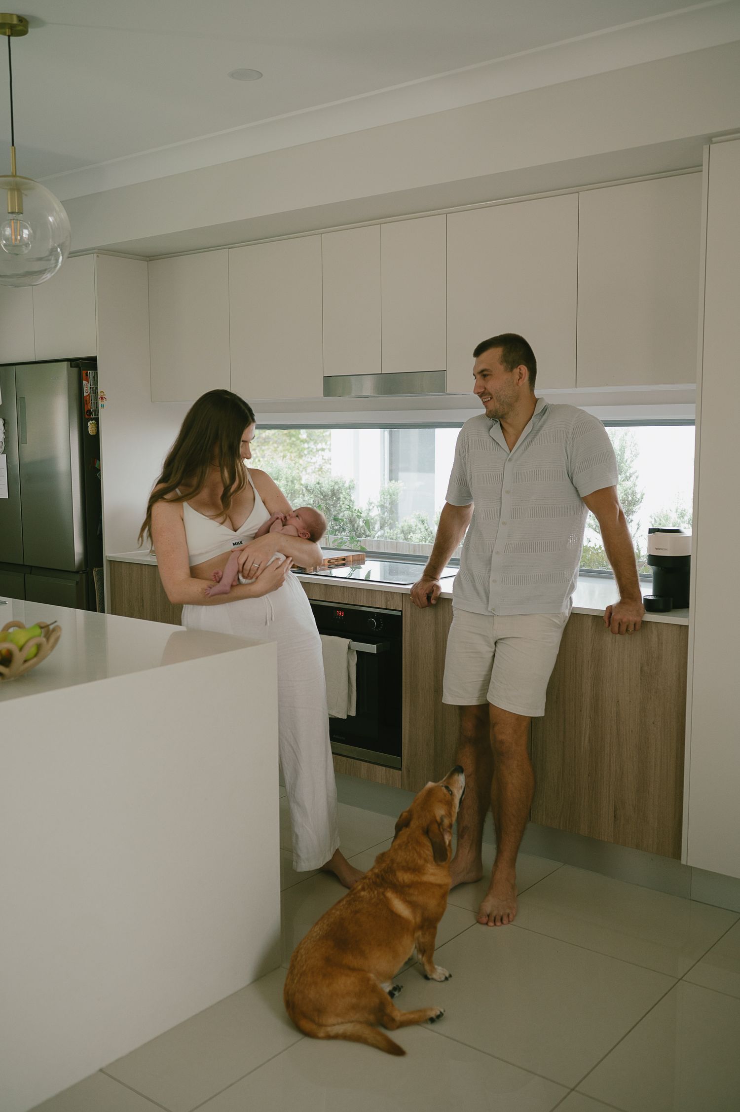 New parents holding newborn baby in modern kitchen with golden dog sitting on floor.