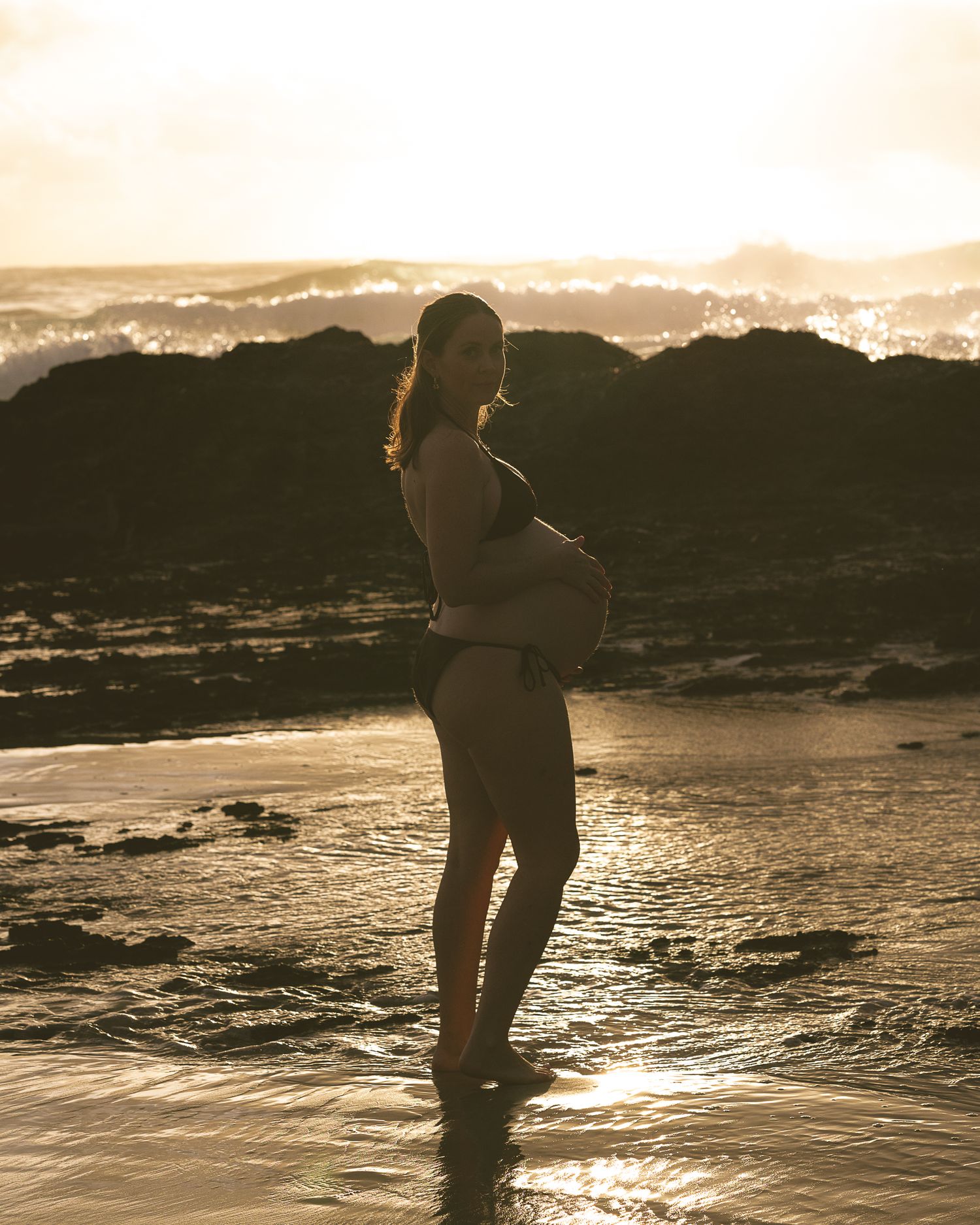 Backlit figure wading in shallow ocean water during golden hour.