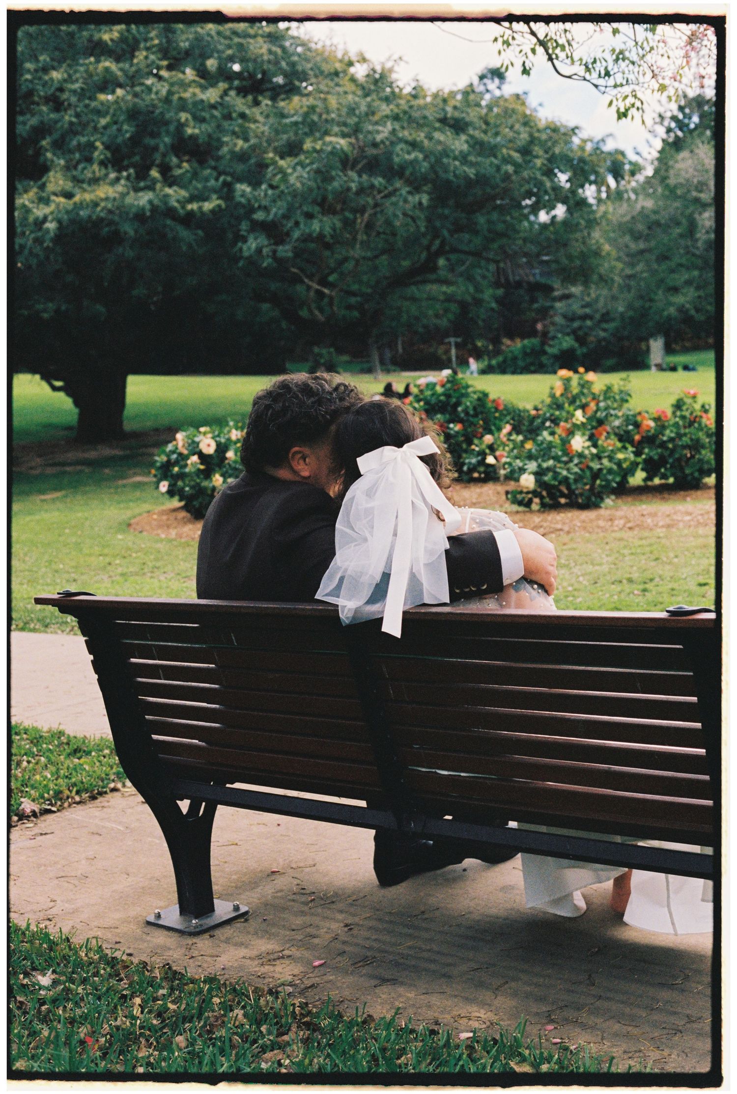 Newlywed couple sharing a romantic kiss on a park bench, bride in white veil and groom in black suit.