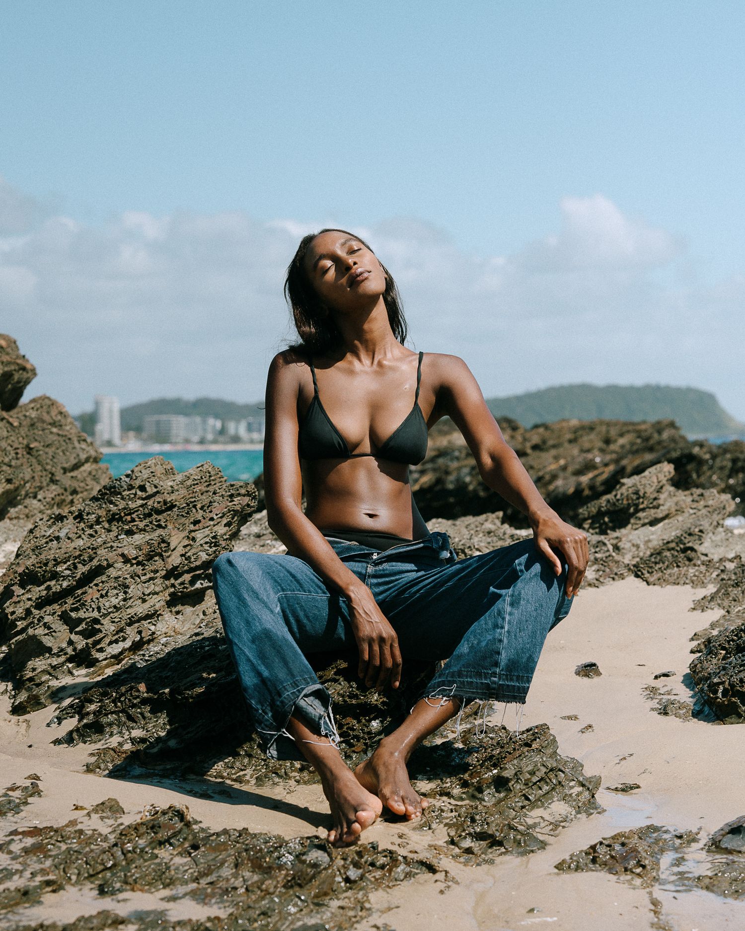 Casual beachwear shoot on rocky beach with city skyline visible in background.