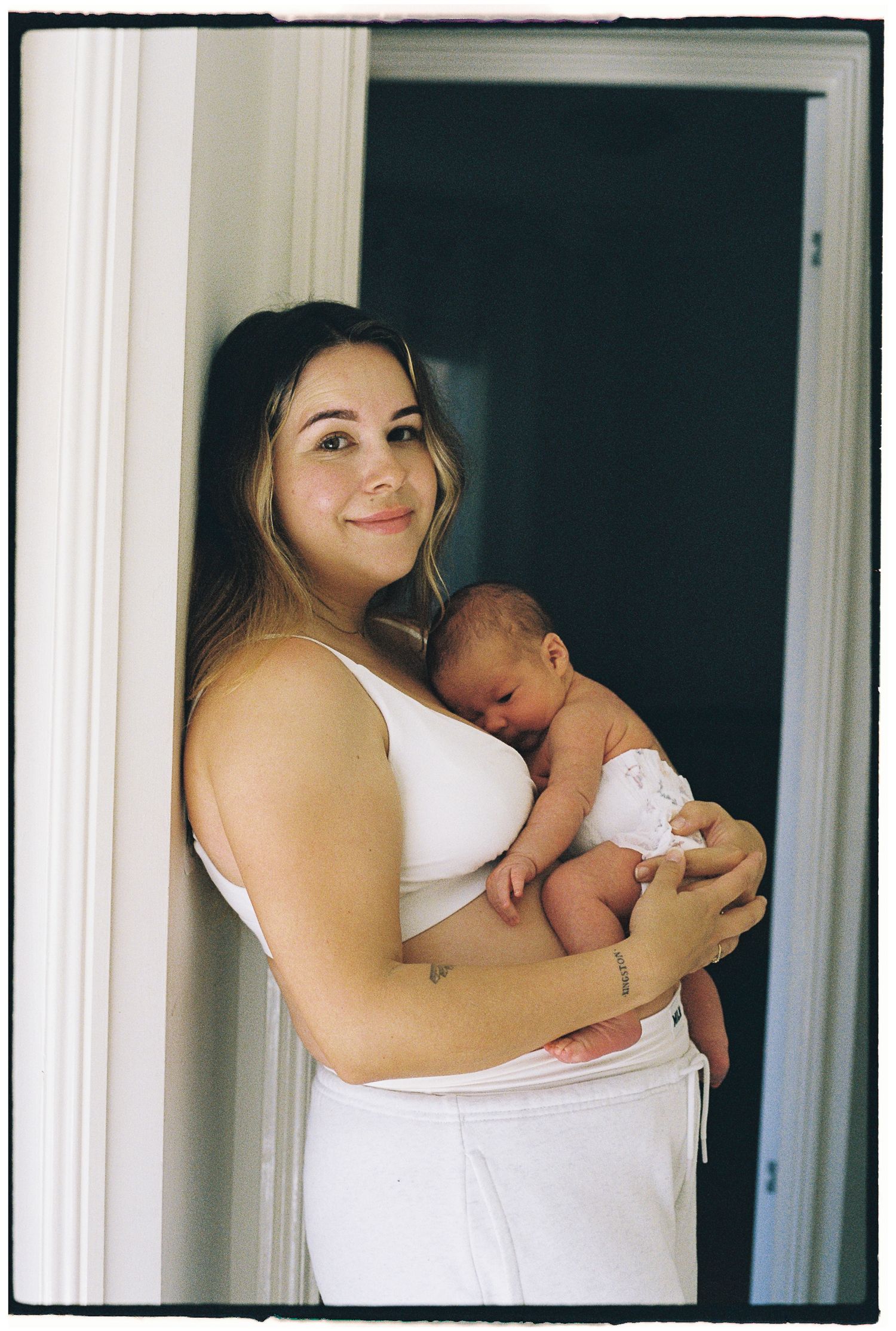 Young mother in white crop top and pants holds sleeping newborn baby while standing in doorway, smiling warmly.