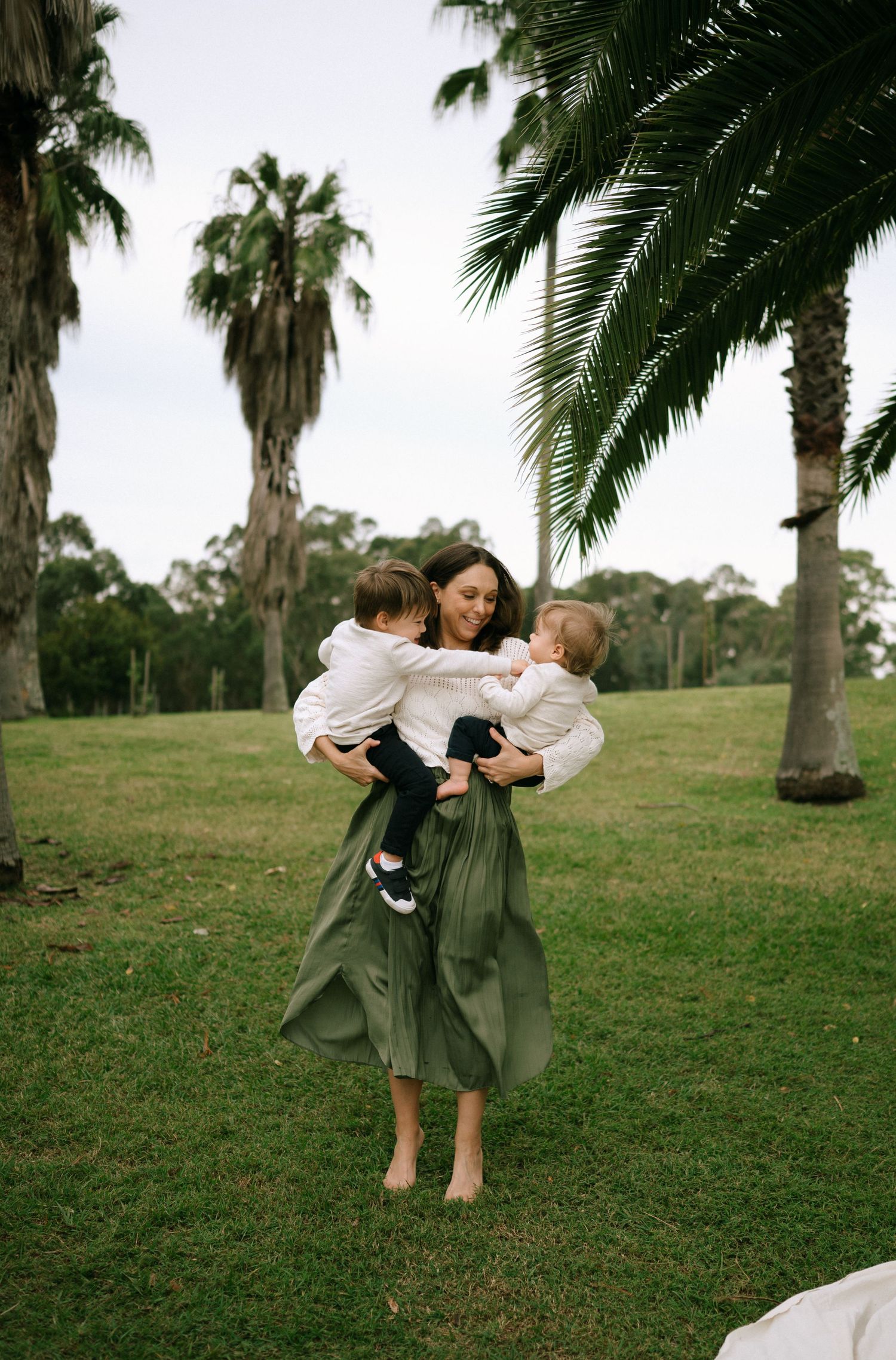 Two figures in matching olive dresses playing together on grassy hill with palm trees.