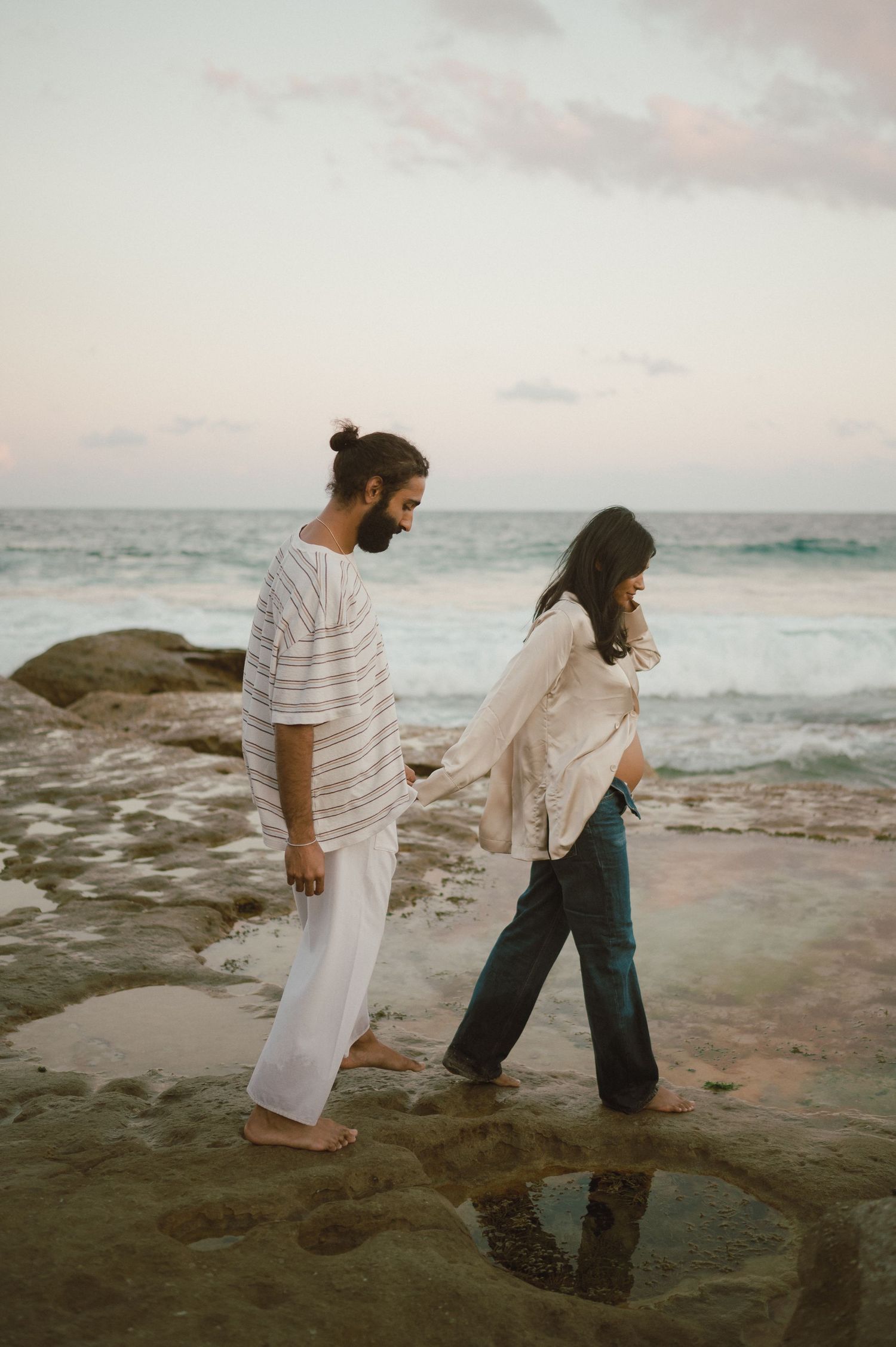 Two individuals walking hand in hand along a rocky shoreline during sunset.