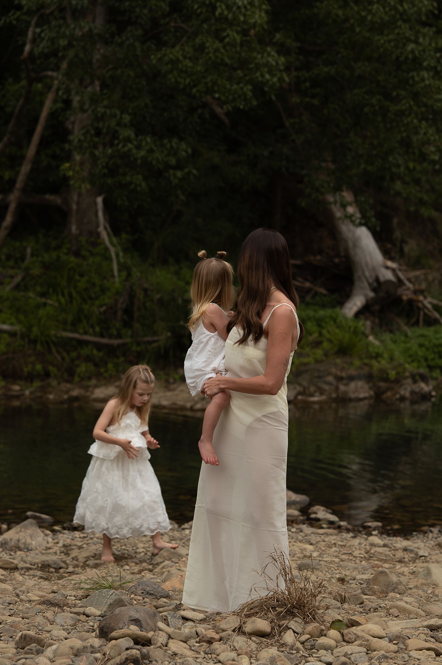 A mother in a white dress holds her toddler by a rocky riverbank while her young daughter in white explores nearby.